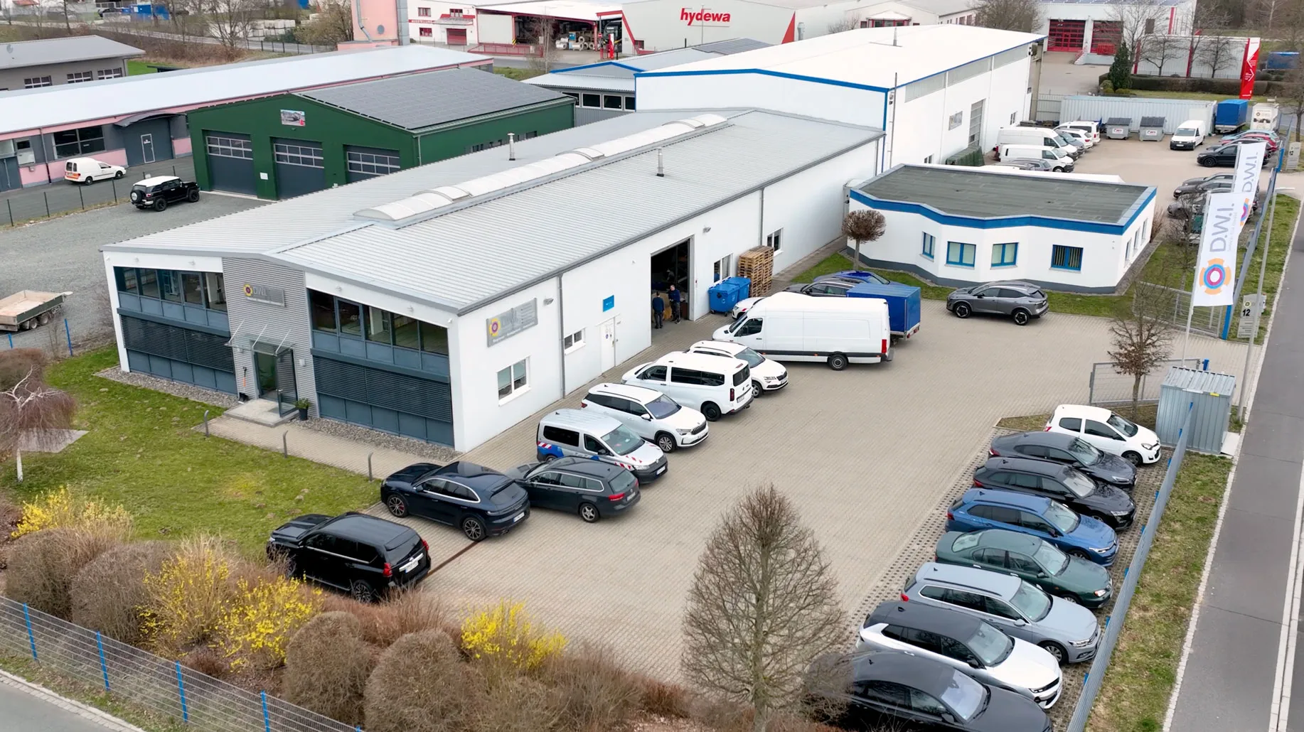 Luftaufnahme eines gewerblichen Gebäudekomplexes mit mehreren geparkten Autos und Firmenflaggen vor dem Eingang.