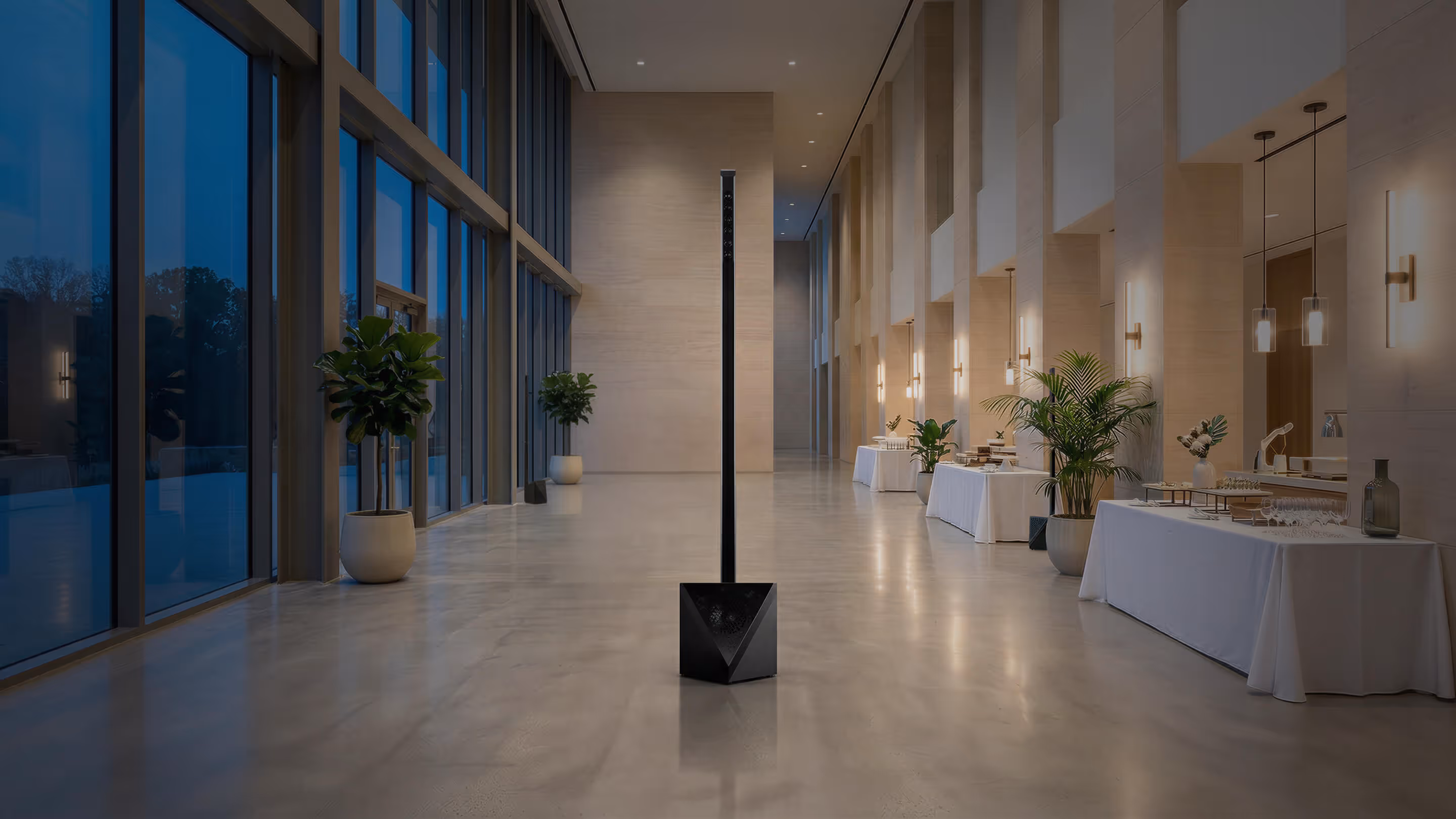 Modern, tall black speaker unit standing in a spacious, well-lit hallway with large windows and tables covered with white cloths and decorations.