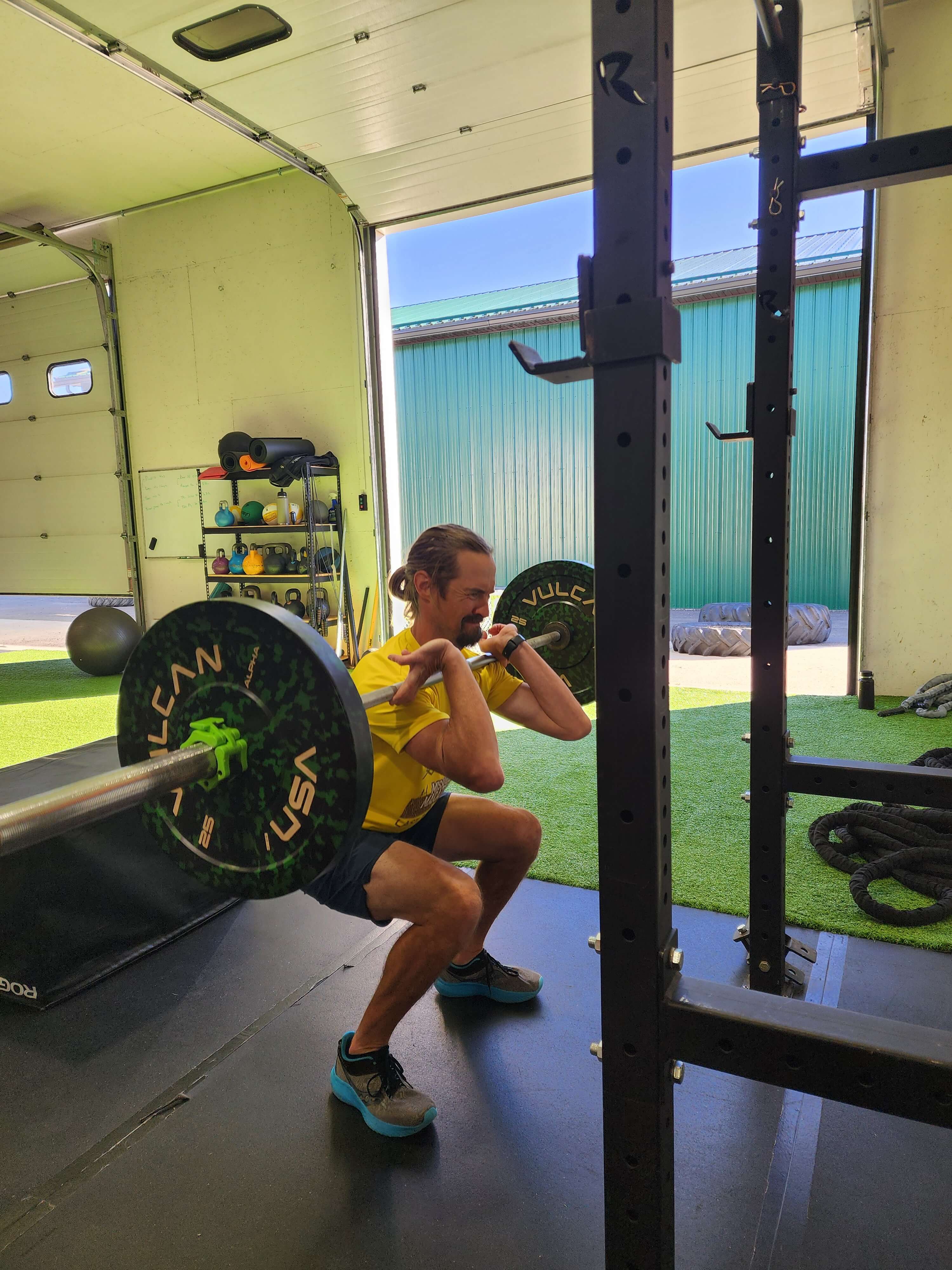 Man in yellow shirt performing a front squat with a barbell in a gym with open doors and exercise equipment in the background.
