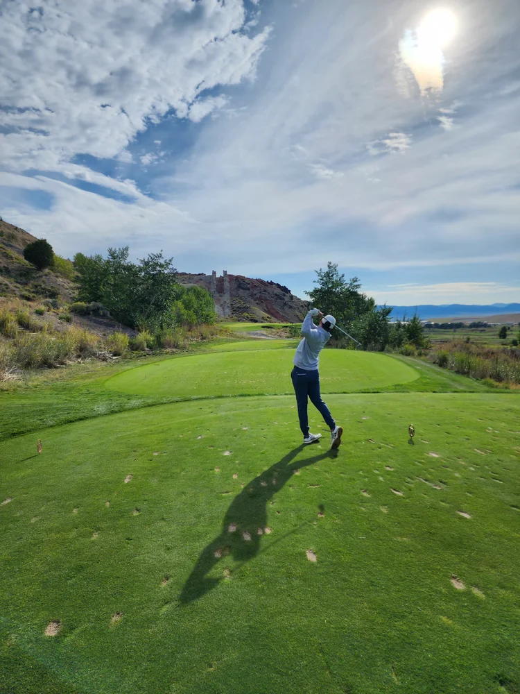Golfer following through on a swing at a green tee with hills, trees, and a partly cloudy sky in the background.
