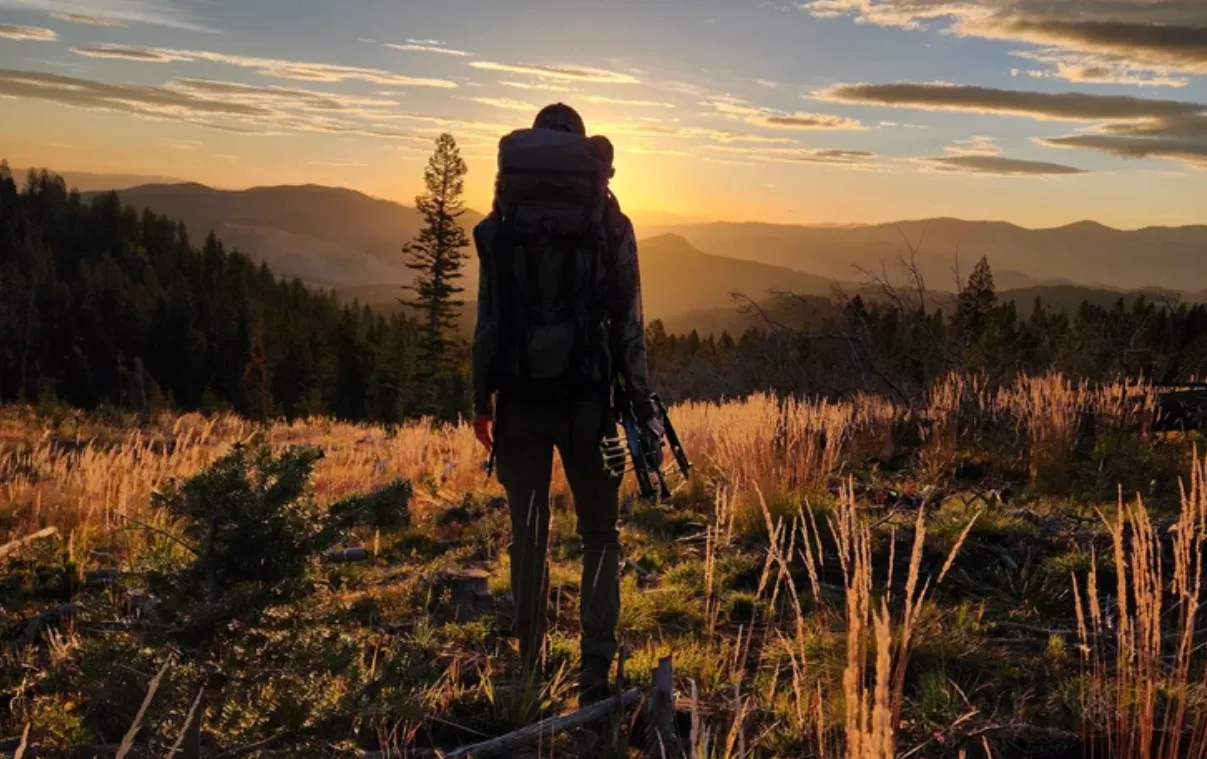 Hiker with backpack and tripod stands facing sunset in a mountainous grassy landscape.
