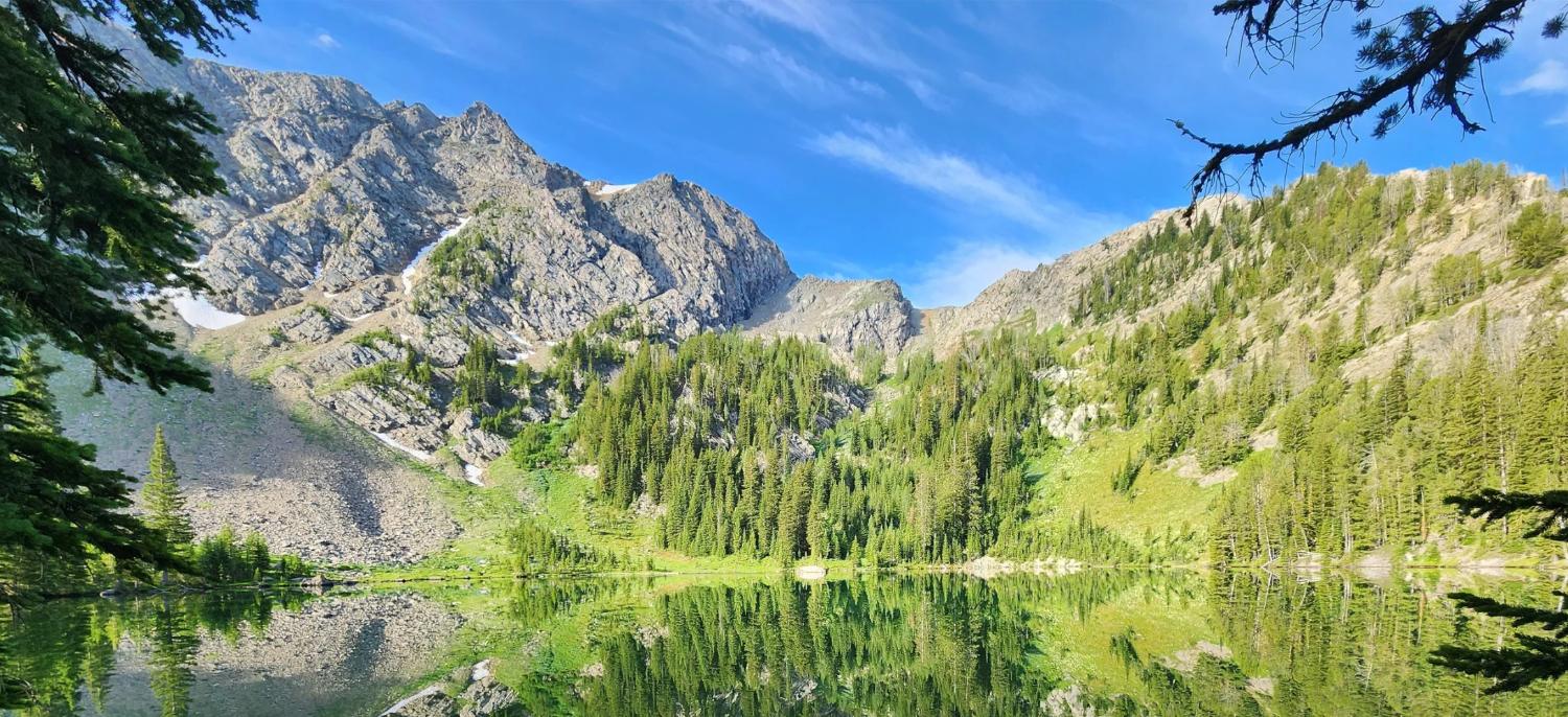 Mountain range with rocky peaks and dense green trees reflected in a clear lake under blue sky.