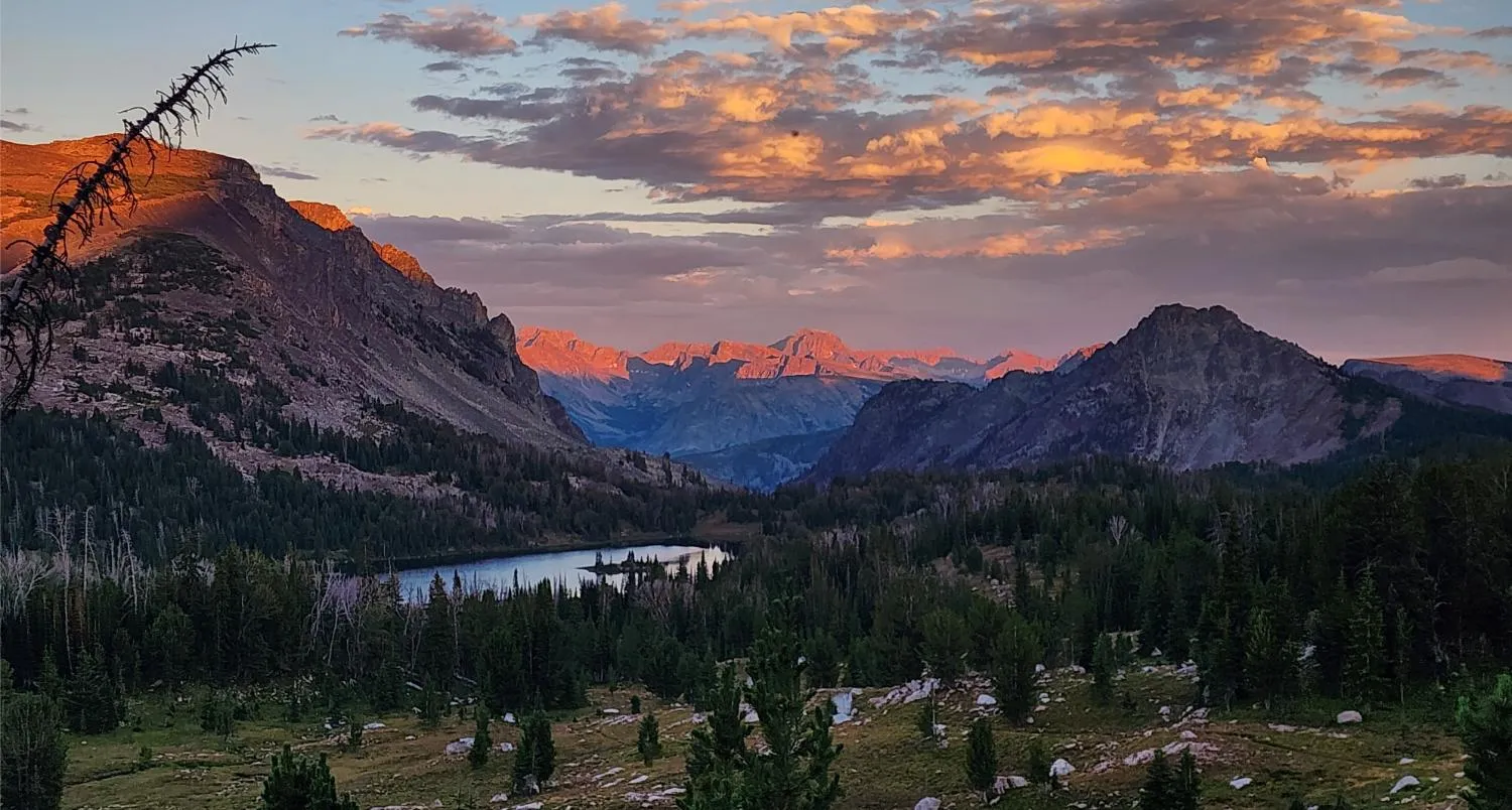 Mountain valley at sunset with forest, a lake reflecting the sky, and clouds lit in orange hues.
