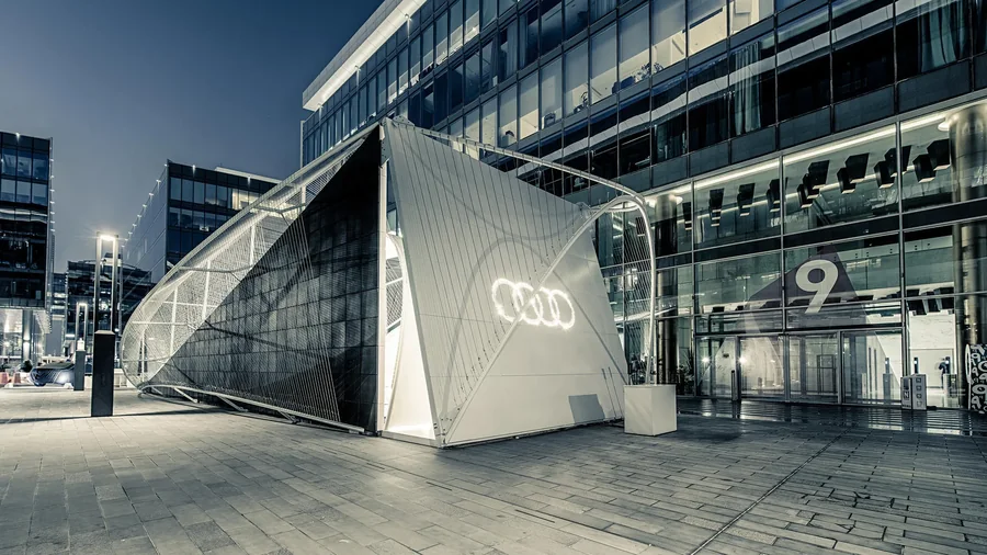 Audi Innovation Hub at night in Dubai Design District: a black rubber-clad side and a white end wall with the lit Audi rings, wrapped in tensile cables.