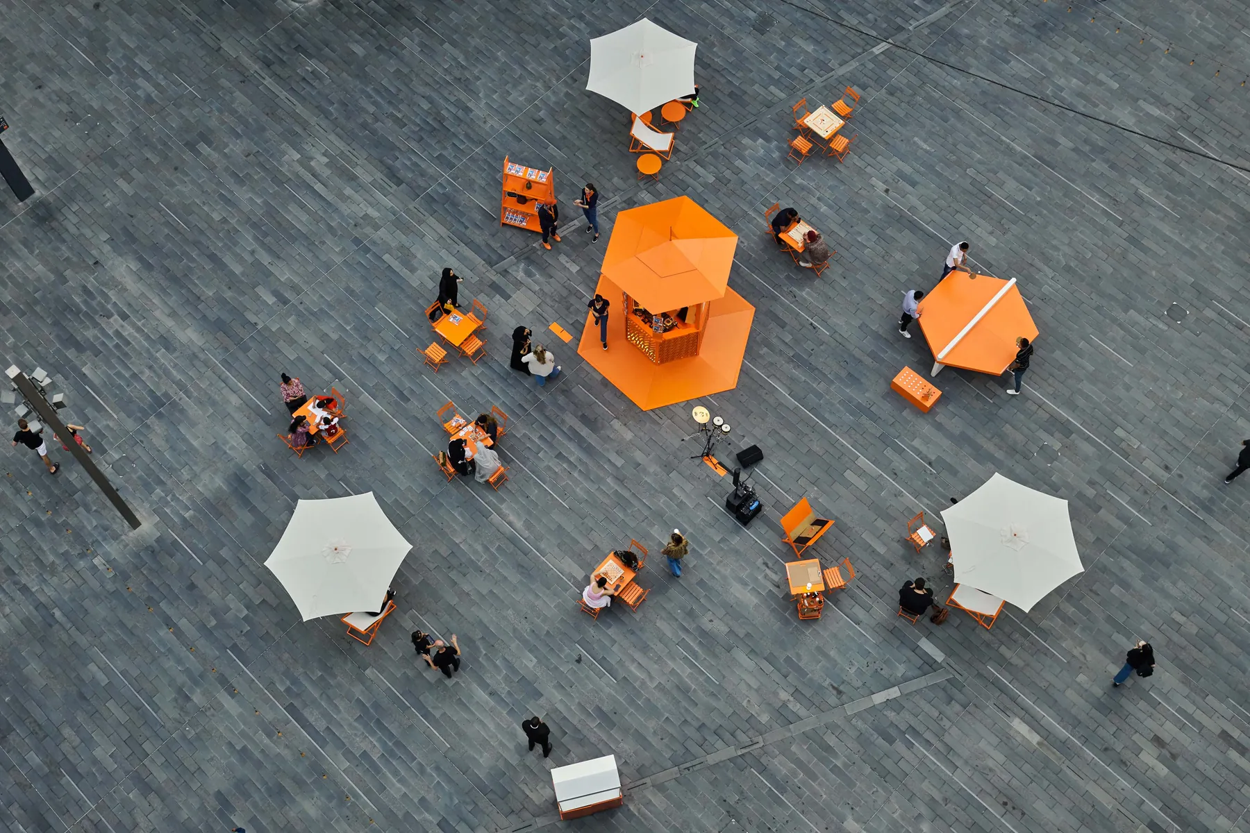 The orange Hermès kiosk seen from above in a paved plaza, with orange tables, chairs and parasols around it.