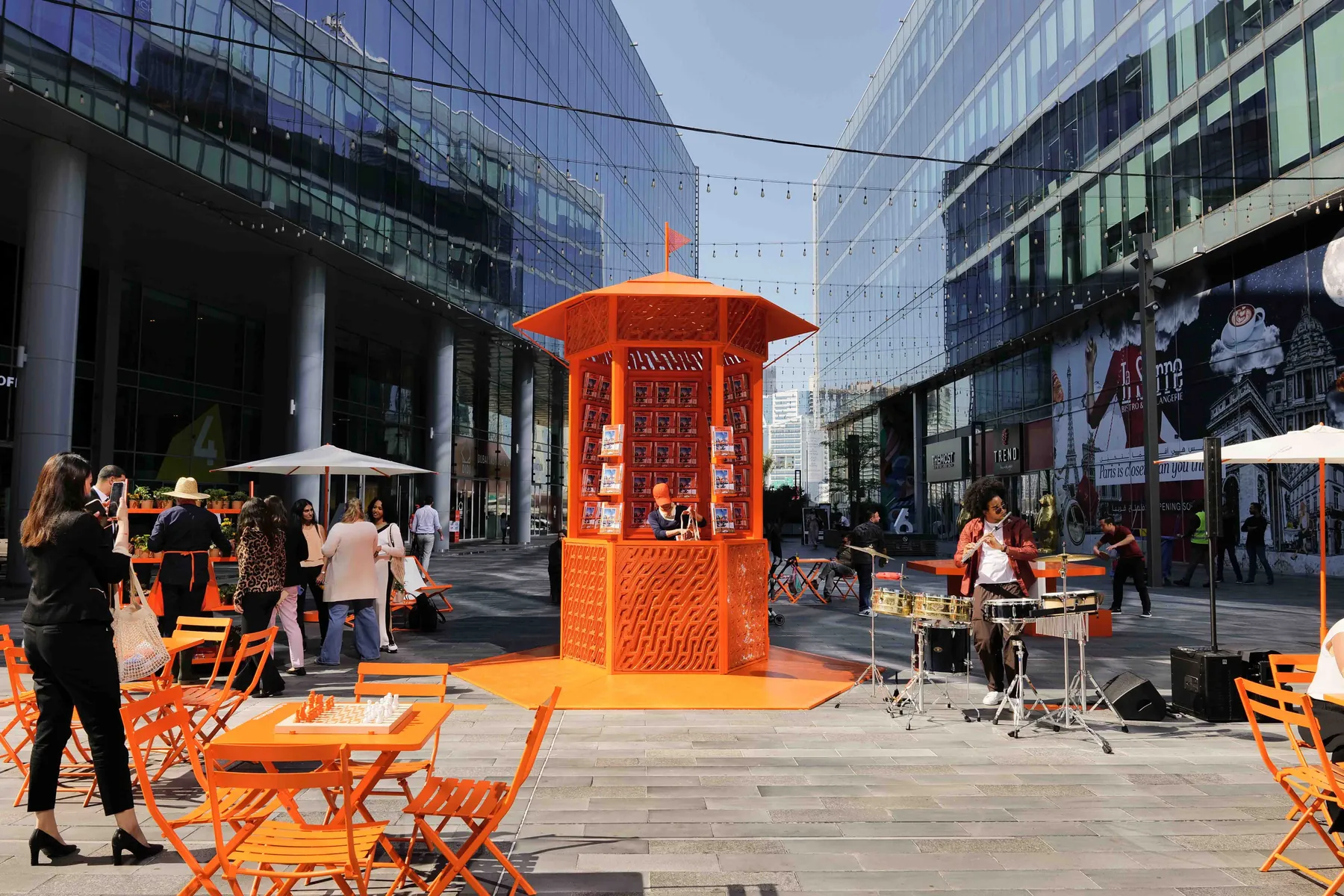 The orange hexagonal kiosk with patterned printed panels in a plaza, people seated around it.