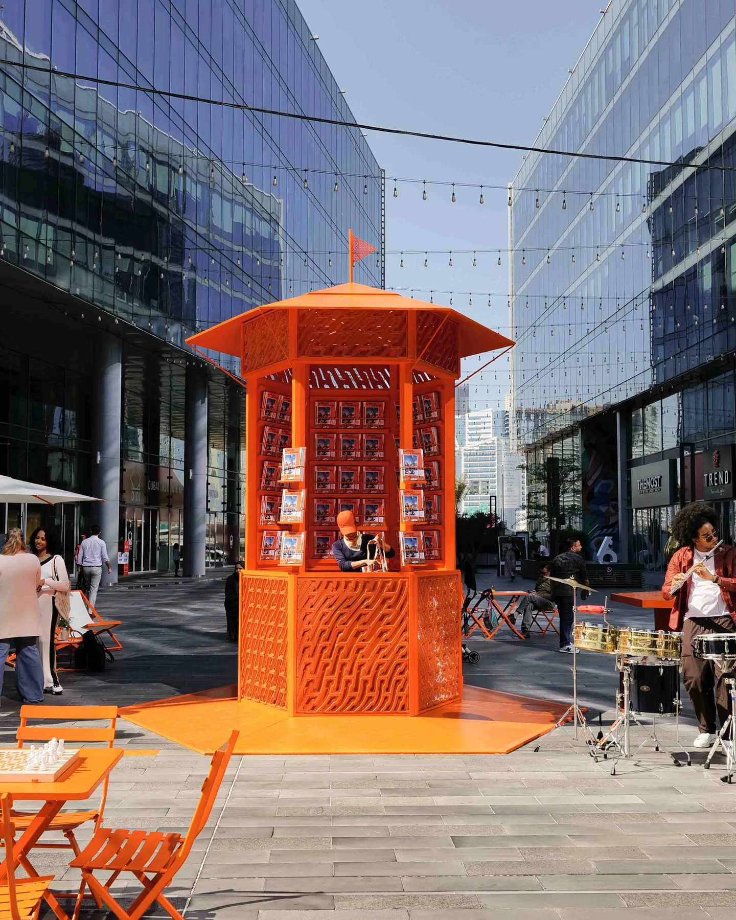 The orange kiosk in Dubai Design District seen square on to its counter, with magazines on display and people around it.