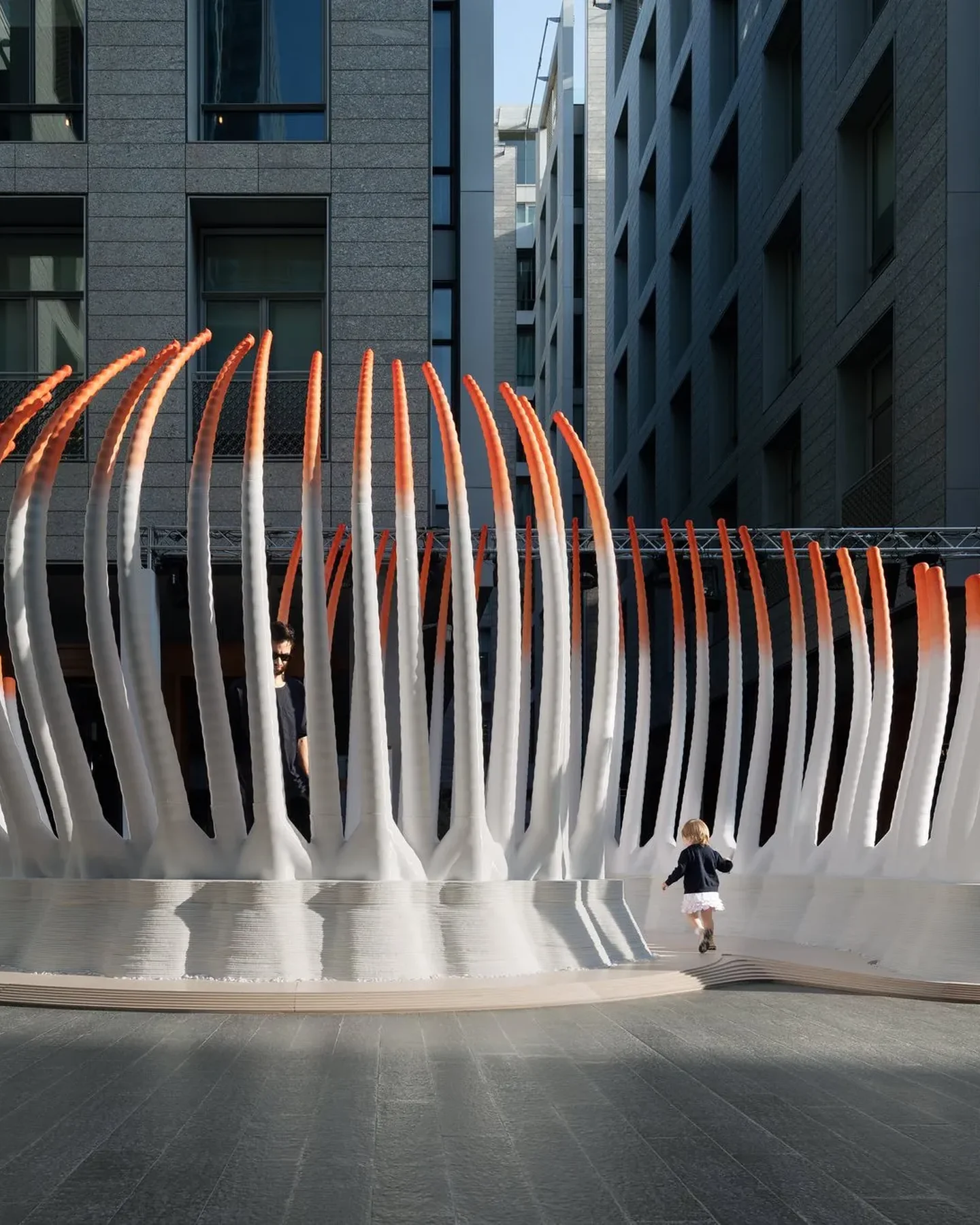 The Deciduous pavilion seen from the side in a DIFC plaza: white 3D printed ribs with orange tips rising from a printed concrete base, with visitors around it.
