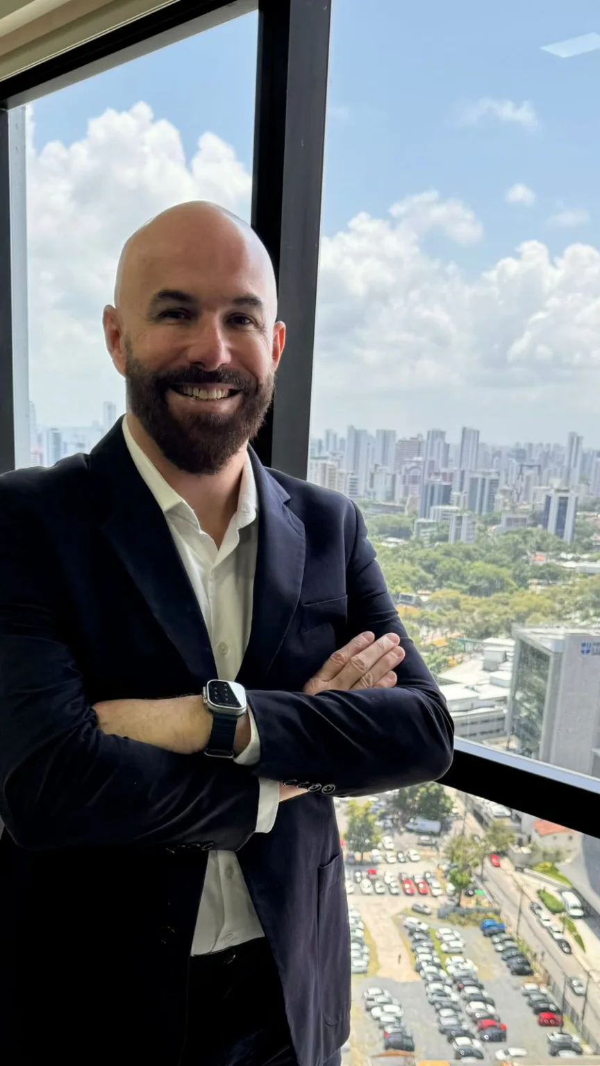 Smiling bald man with beard in black blazer stands with arms crossed by a window with city view.