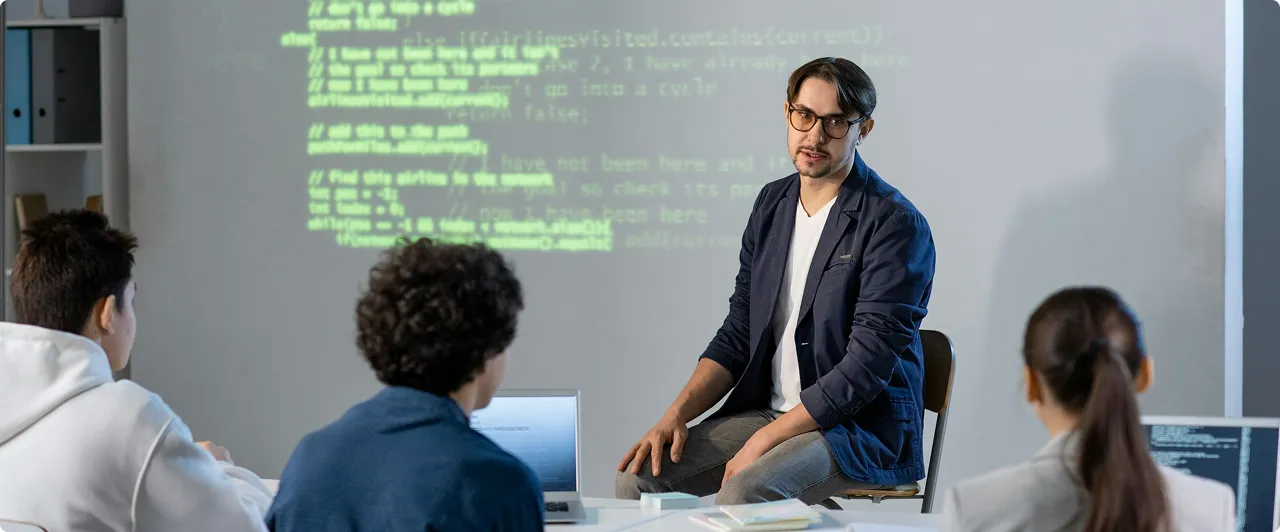 Un homme en veste assis face à trois personnes écoutant une présentation avec du code projeté sur un mur.