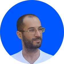 Portrait d'un homme souriant avec des lunettes et une barbe, portant une chemise blanche avec les bras croisés sur un fond bleu.