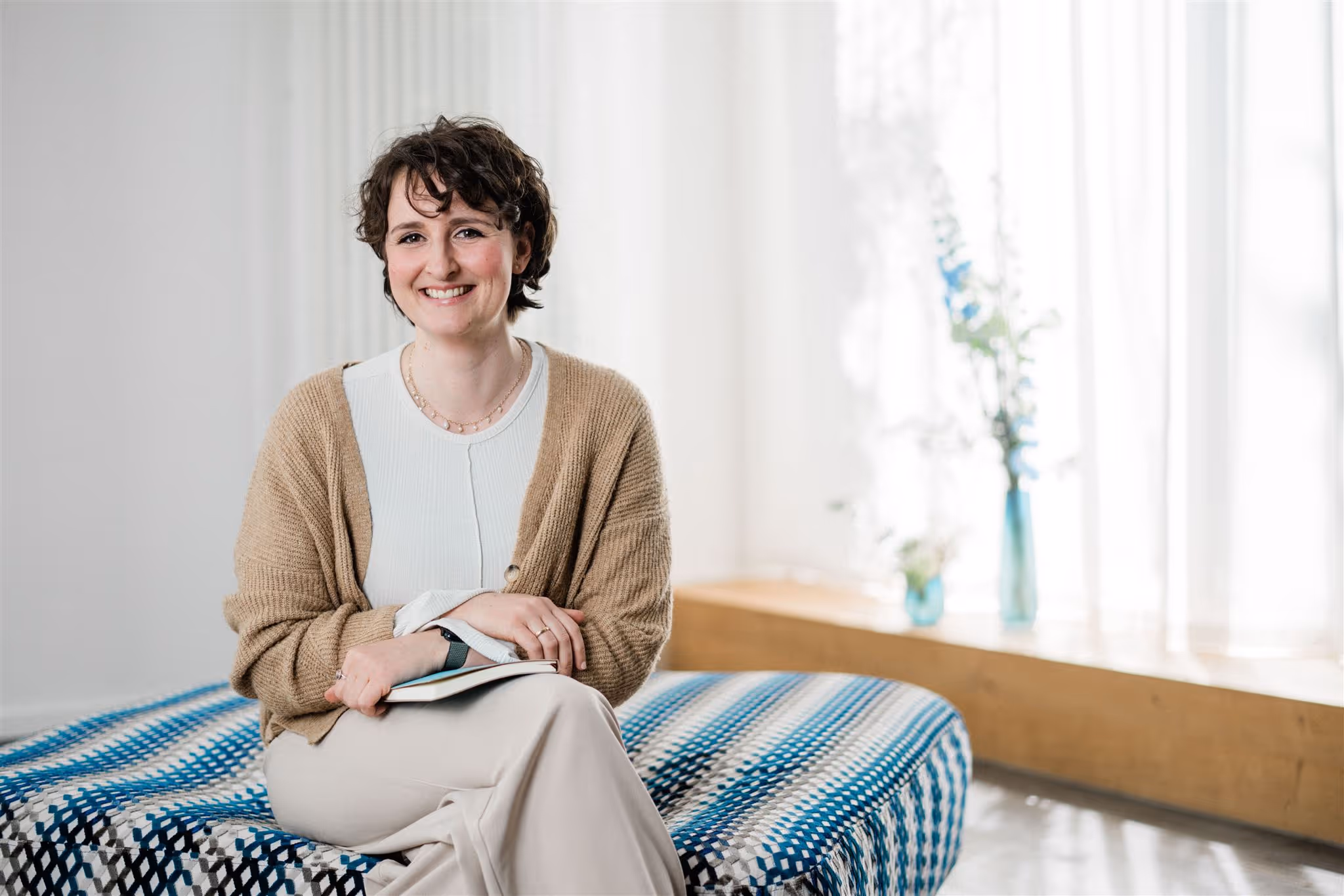 Julia Bobe mit beiger Strickjacke und weißem Oberteil sitzt auf einer gemusterten Decke und hält ein Buch.