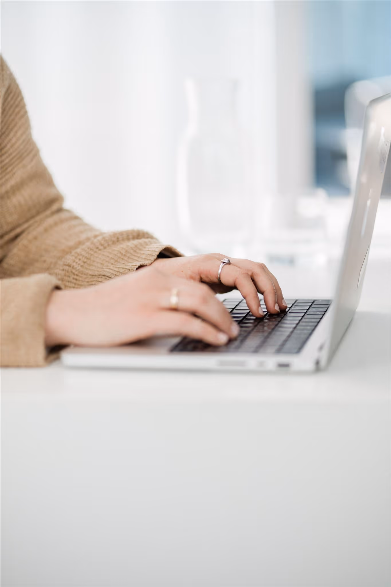 Person mit Ringen tippt auf Tastatur eines silbernen Laptops an einem weißen Tisch.