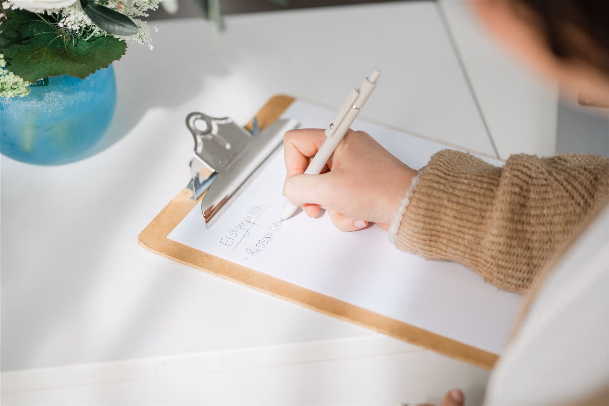 Eine Person schreibt mit einem weißen Stift auf einem Klemmbrett, auf dem Papier steht das Wort 'Ersparniss' und 'Ressourcen'.