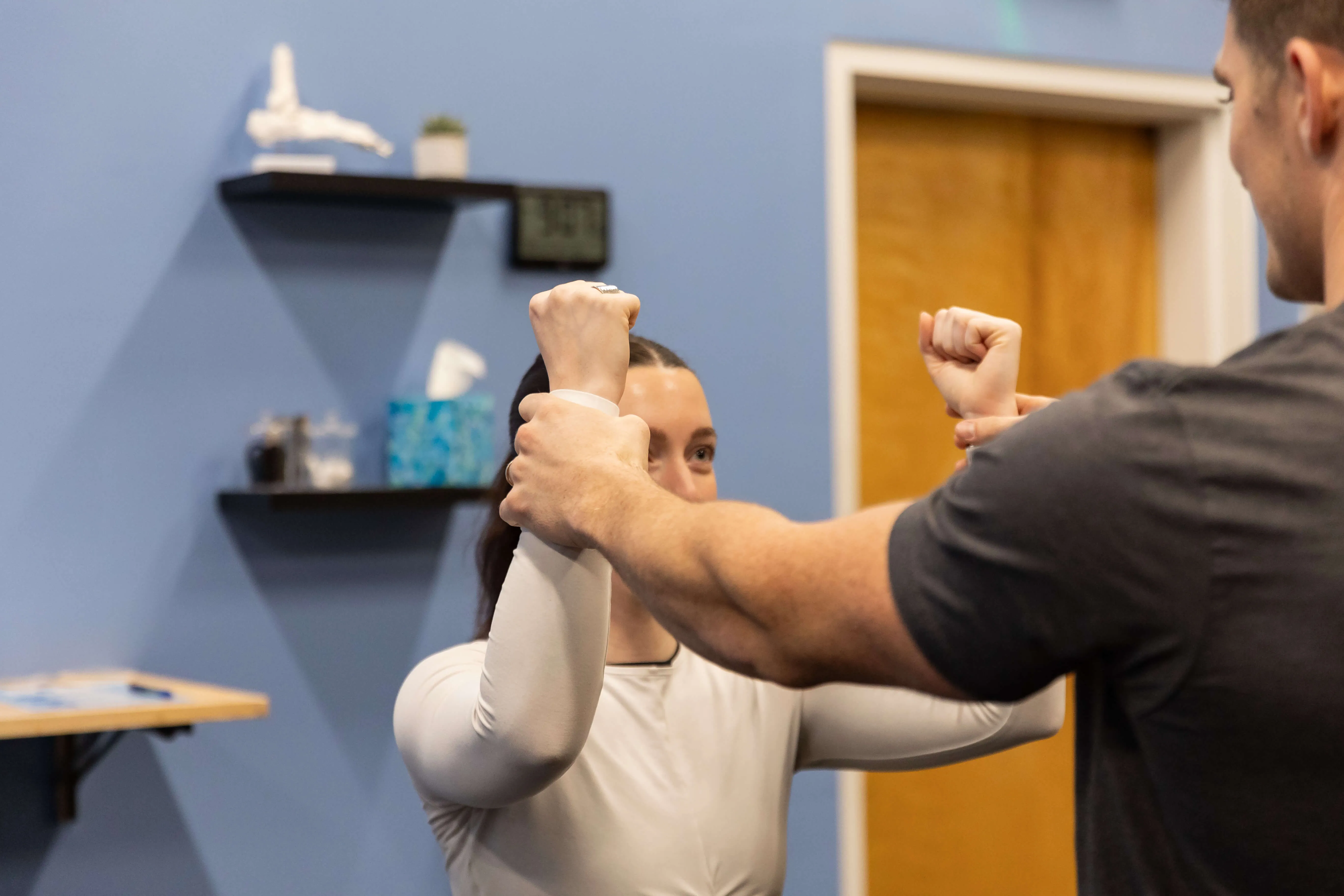 Woman performing arm exercise with assistance from a man in a room with blue walls and shelves in the background.