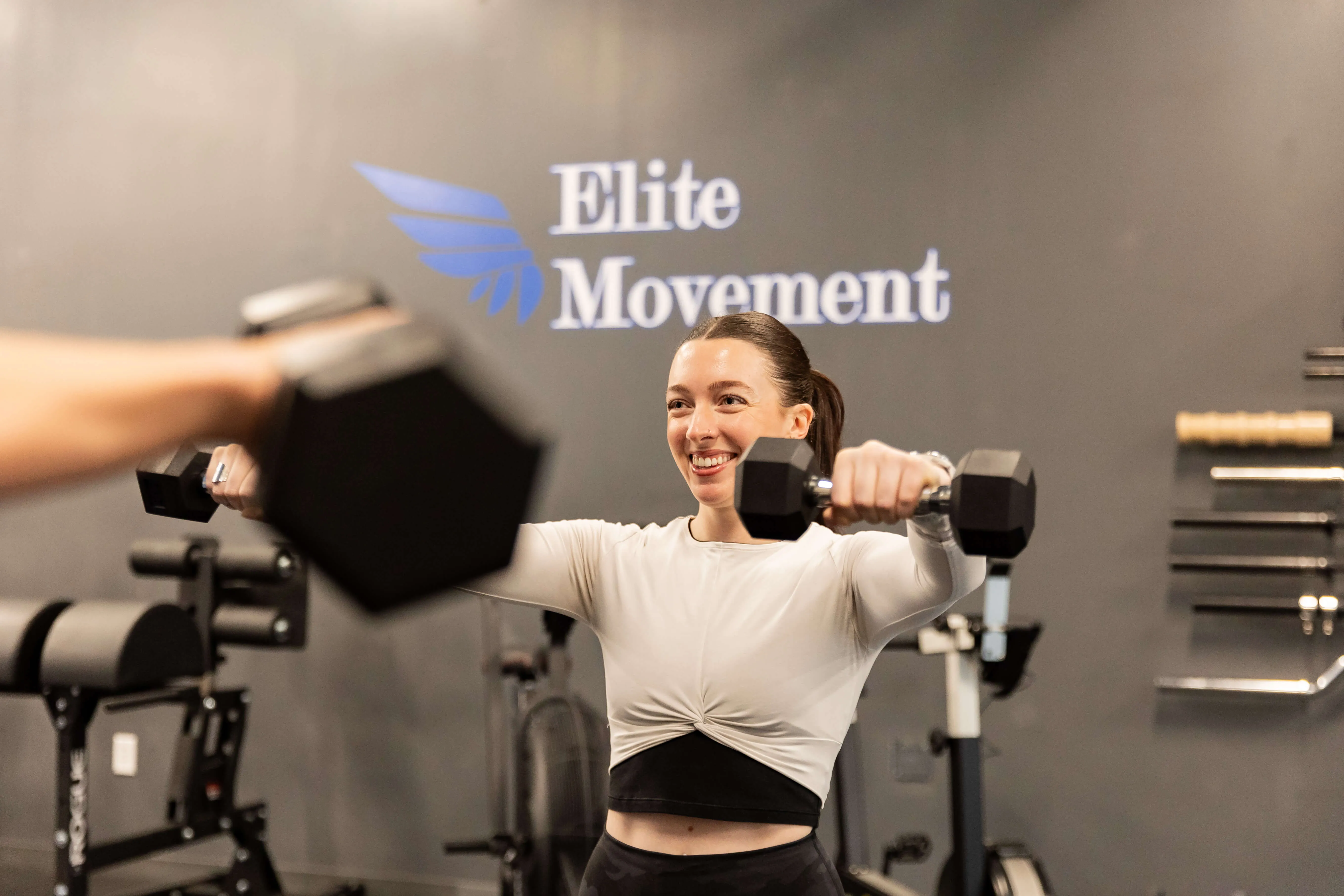 Smiling woman lifting dumbbells in a gym with Elite Movement logo on the wall behind her.