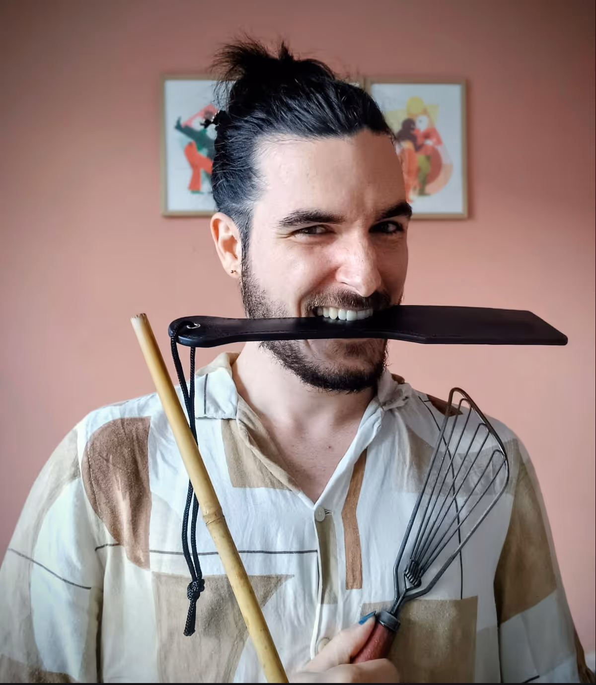 Man with mustache and beard holding kitchen tools, biting a black spatula, smiling at camera.
