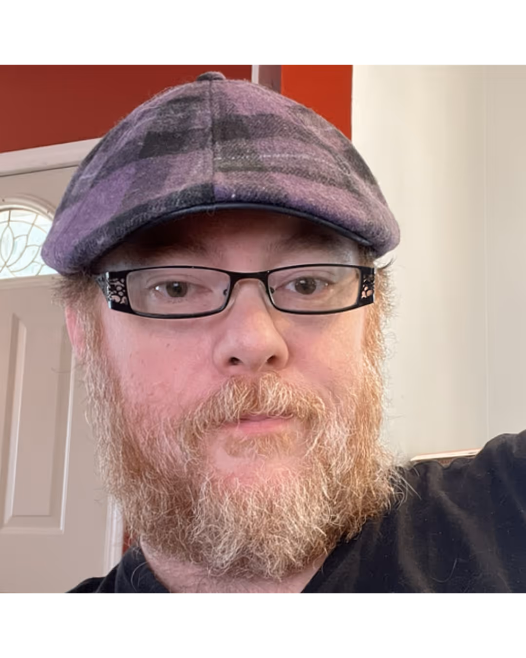 Close-up of a man with glasses, a patterned cap, and a full beard indoors near a door.