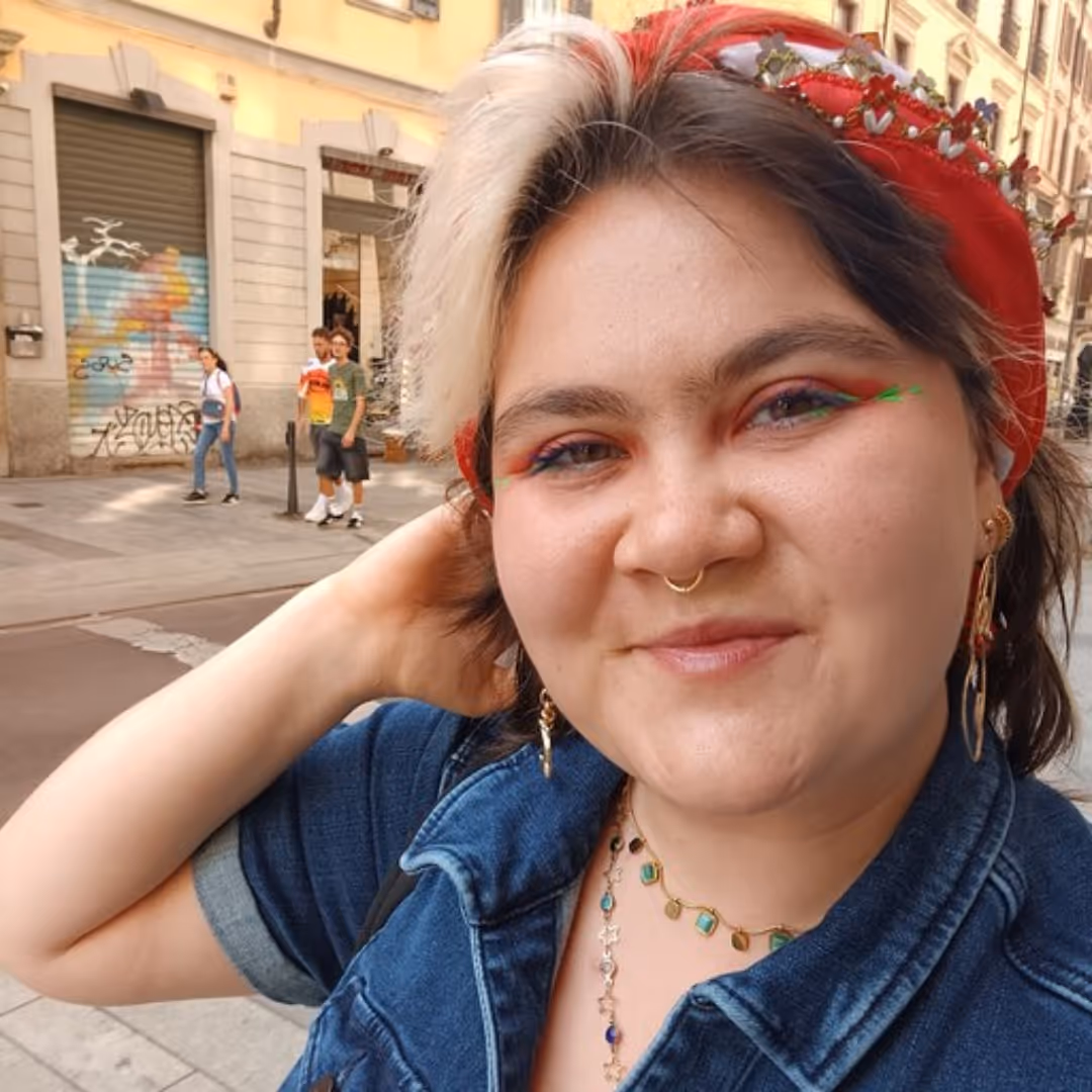 Person with half-white half-brown hair, red headband, colorful eye makeup, and denim jacket on city street.