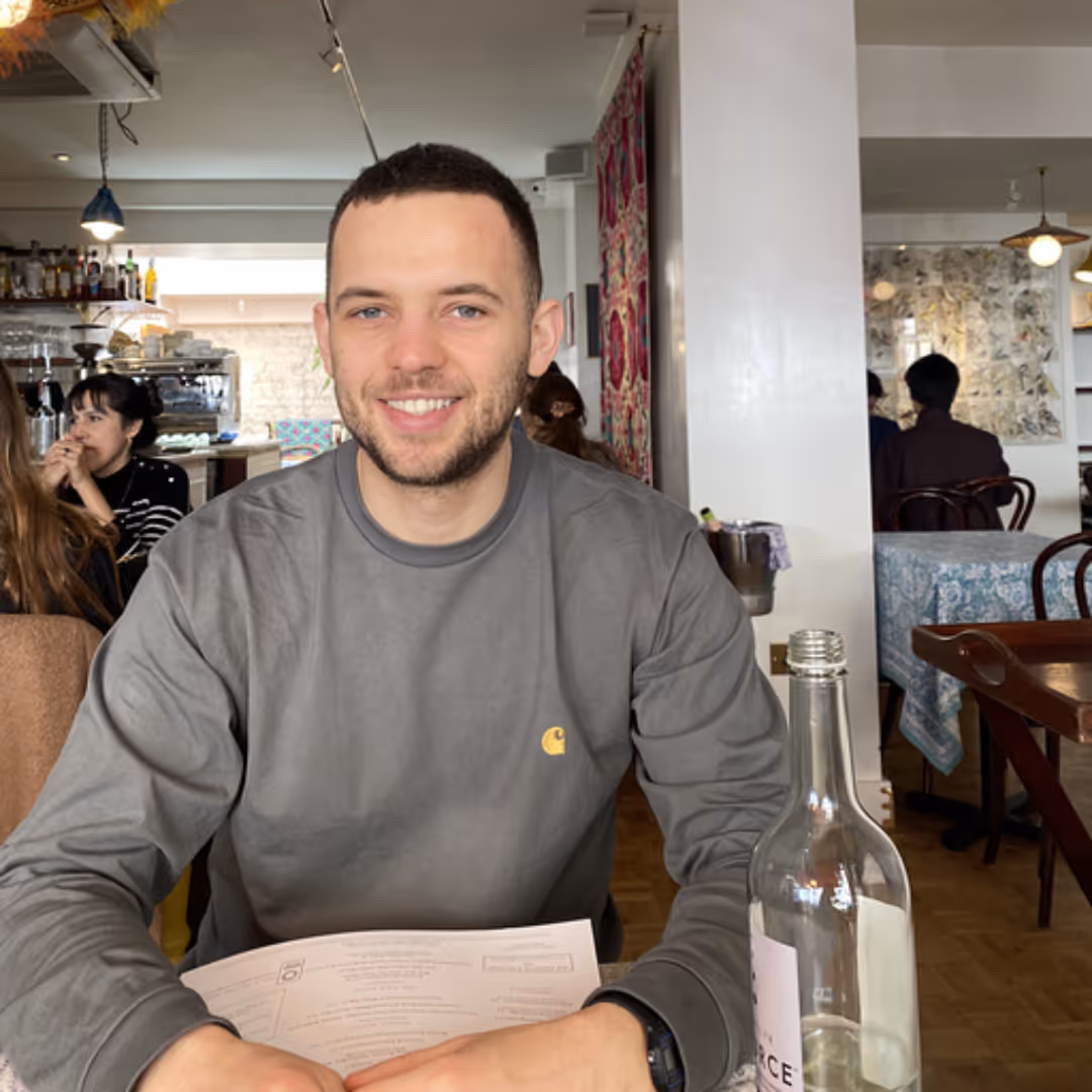 Smiling man in gray shirt holding a menu, seated at a table in a café with a water bottle in front.