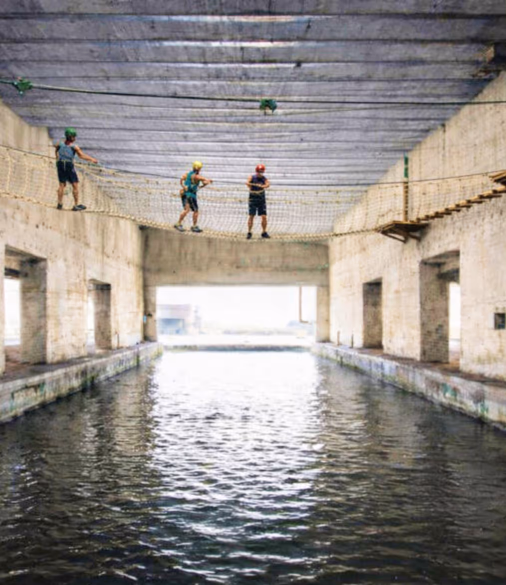 Three people wearing helmets walking on a rope net bridge suspended over water inside a concrete structure.