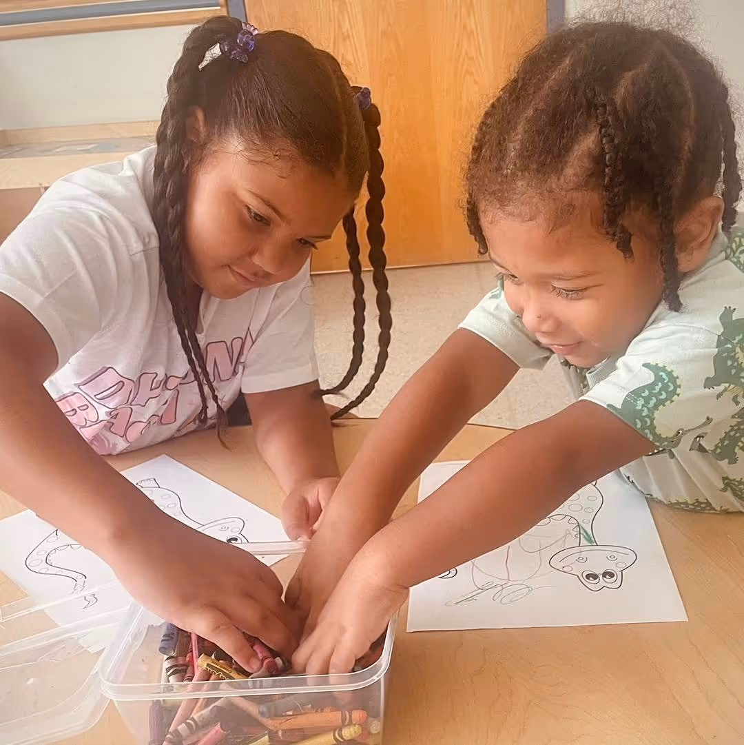 Two children sitting at a classroom table working together on coloring activities, selecting crayons and focusing on a shared task in a supportive learning environment