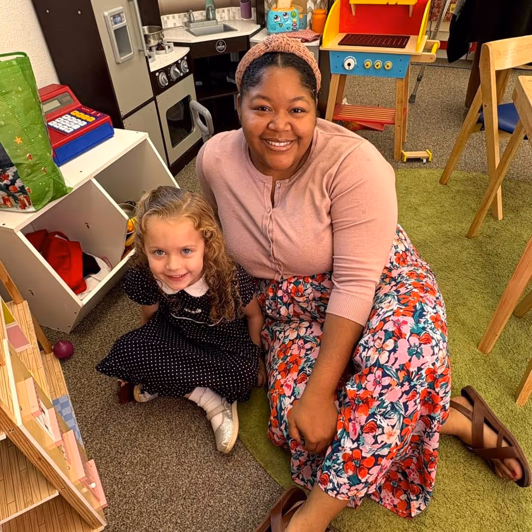 eacher sitting on the classroom floor with a young student, both smiling and engaged in a supportive learning environment surrounded by educational toys and materials