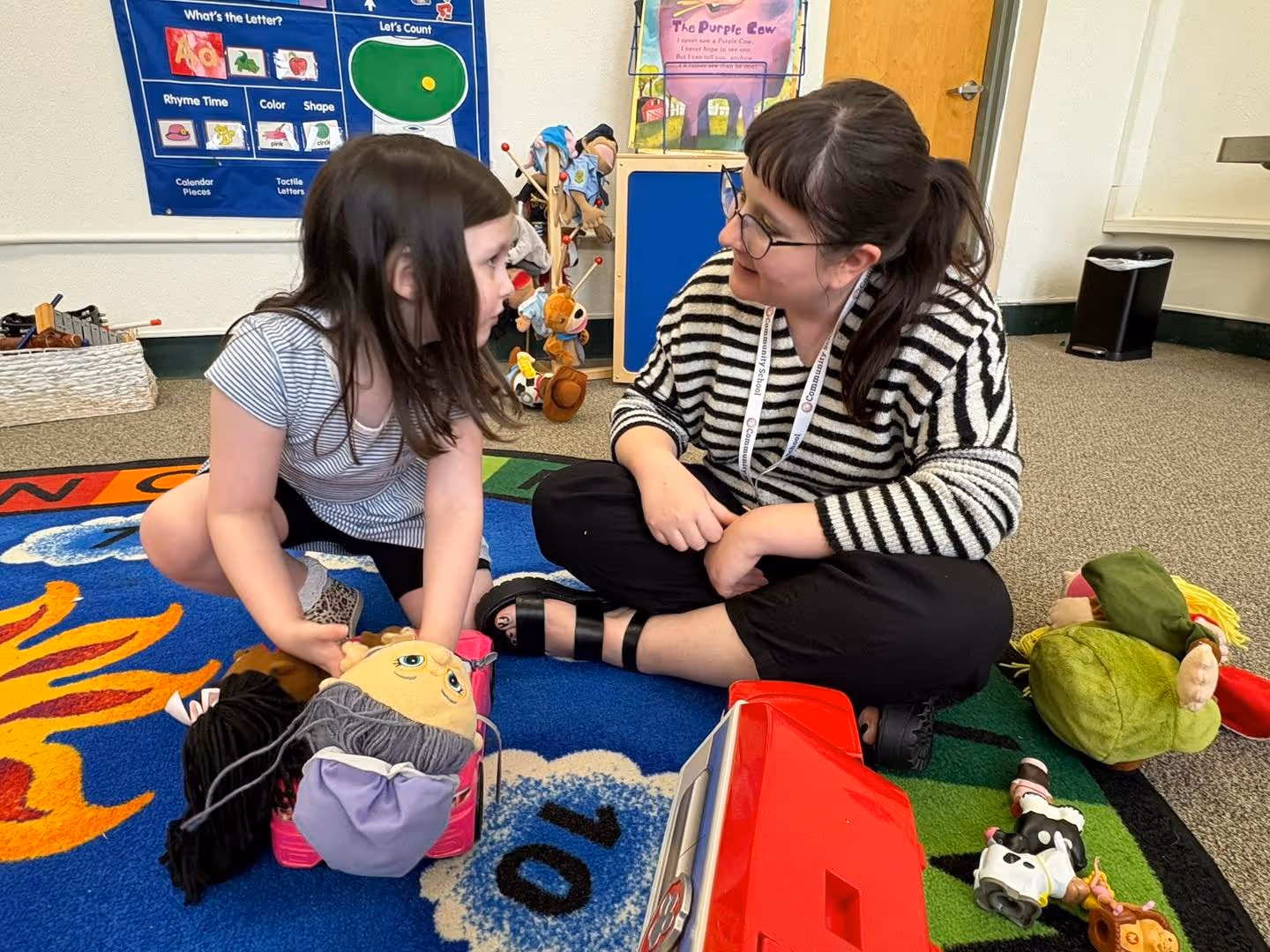 Child sitting on a classroom rug engaging in a one-on-one speech therapy session with a therapist, using toys and visual materials to support communication and interaction skills in a supportive learning environment
