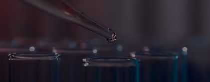 Close-up of a dropper releasing a liquid droplet above test tubes in a dimly lit laboratory setting.