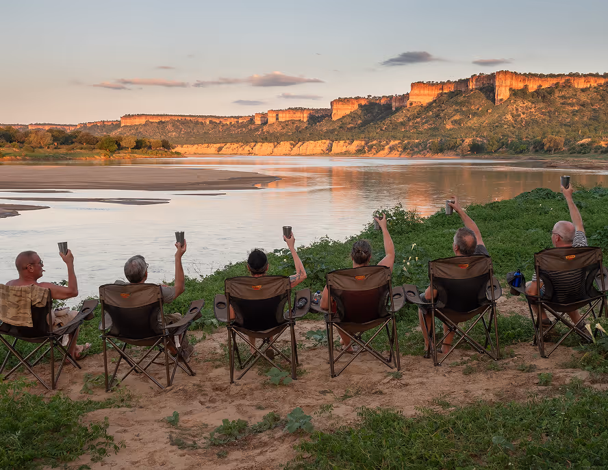 Zes mensen zitten in campingstoelen aan een rivierbank met opgestoken bekers, uitkijkend op rotsachtige kliffen in de avondzon.