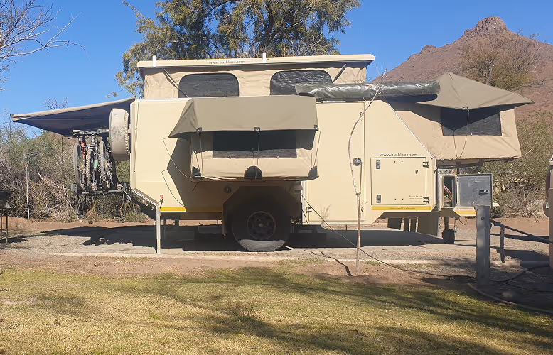 Beige safari kampeerwagen met uitgeklapte luifels, fietsenrek en uitklapbare slaapgedeelten in een droge, bergachtige omgeving.