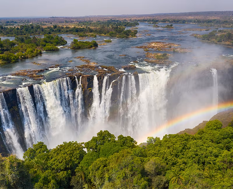 Grote waterval met een regenboog en dichtbegroeid groen rondom onder een blauwe lucht.