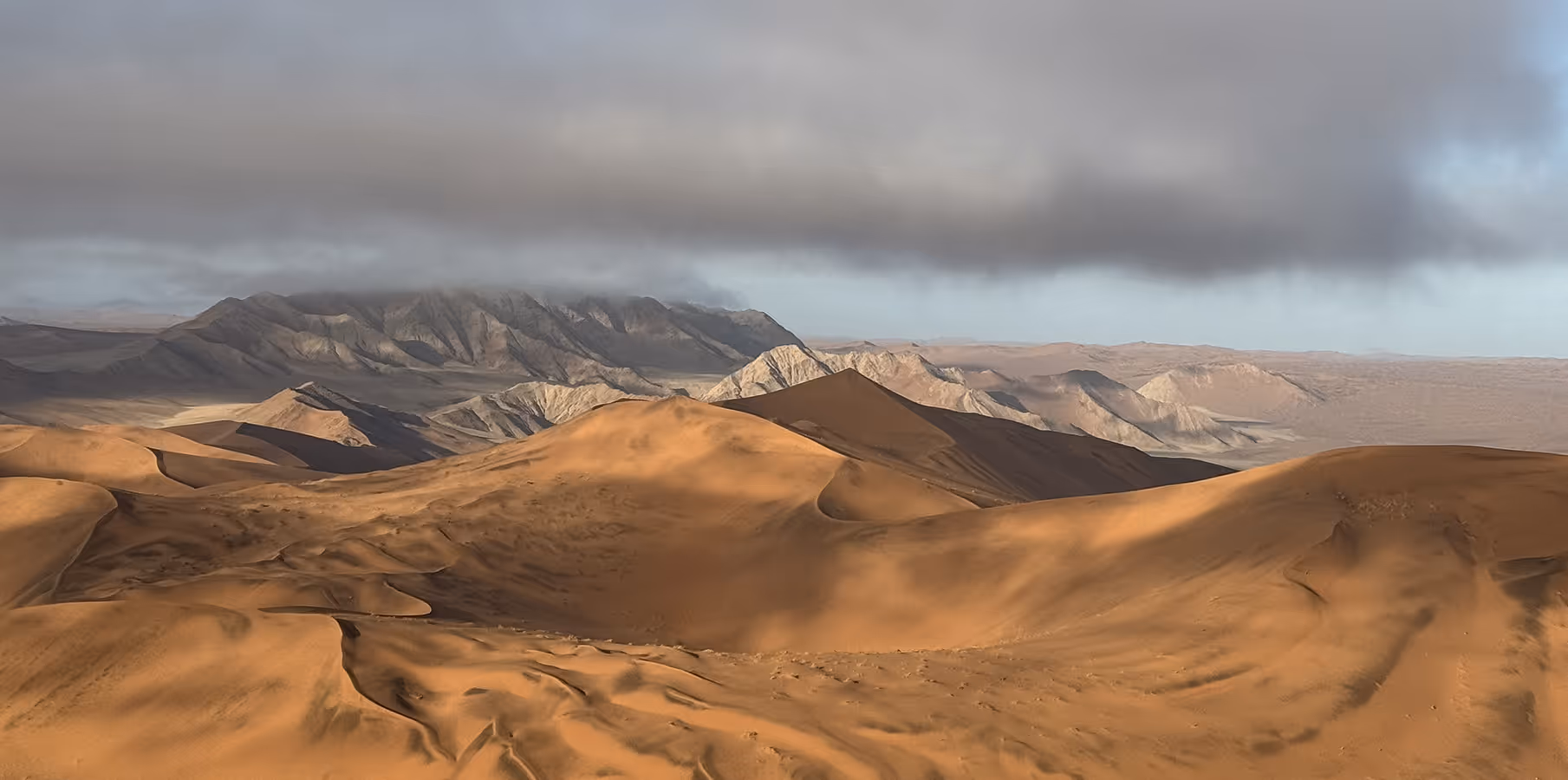Landschap van Namibische zandduinen met bergen in de achtergrond onder een bewolkte hemel.