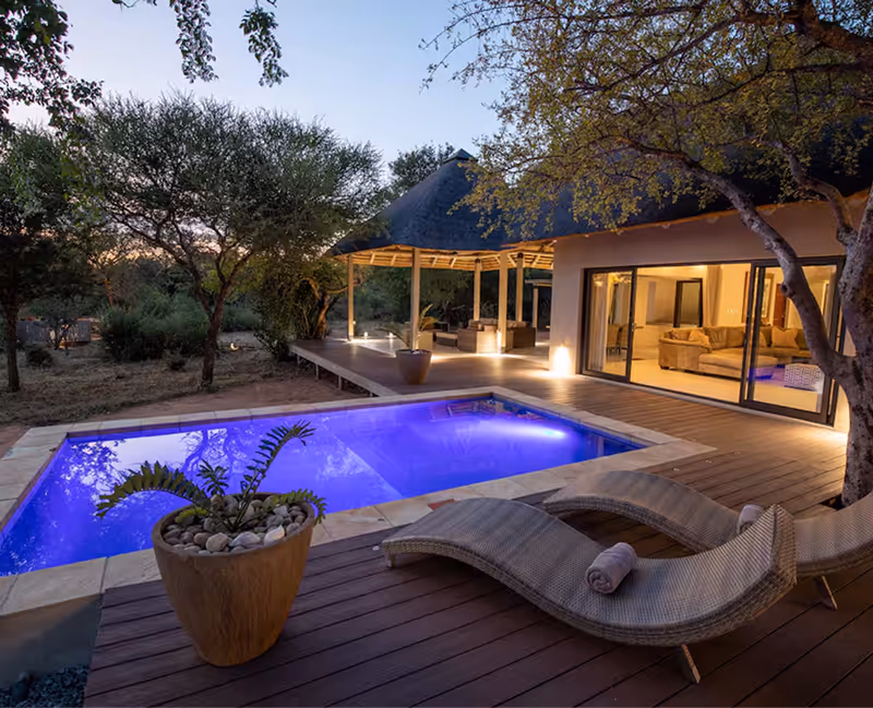Verlichte zwembad bij Villa Muningi met houten terras, ligstoelen en een glazen schuifdeur naar een woonkamer, omgeven door bomen bij schemering.