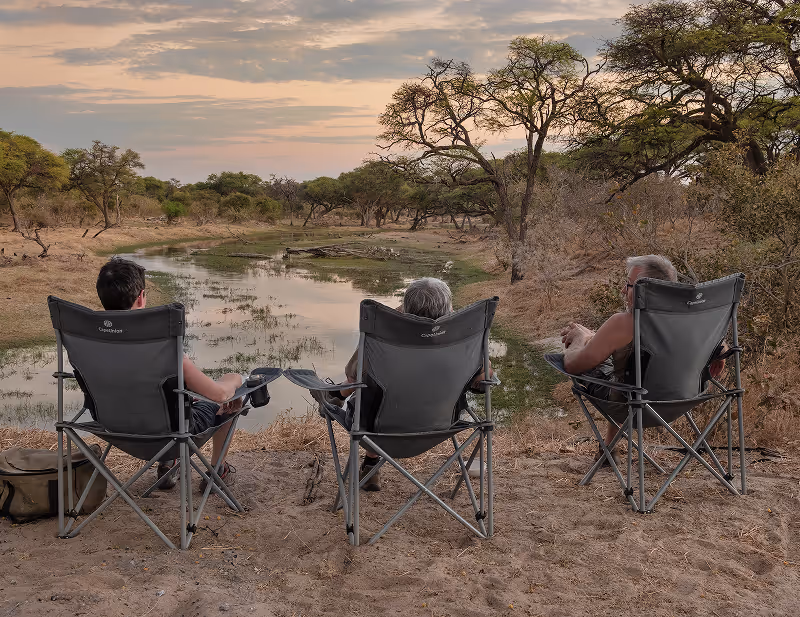 Drie mensen zitten in inklapbare stoelen aan de rand van een rivier in een droge, bosrijke omgeving tijdens zonsondergang.