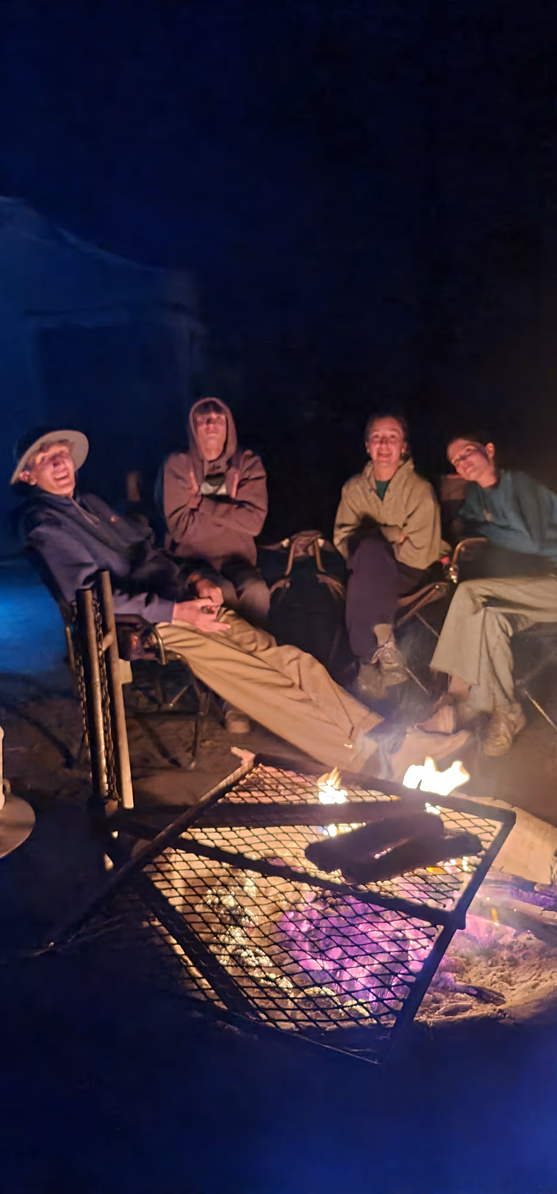 Vier mensen ontspannen rond een kampvuur in de nacht, zittend op campingstoelen met gloeiende houtskool en brandend vuur in het vuurkorfrek.