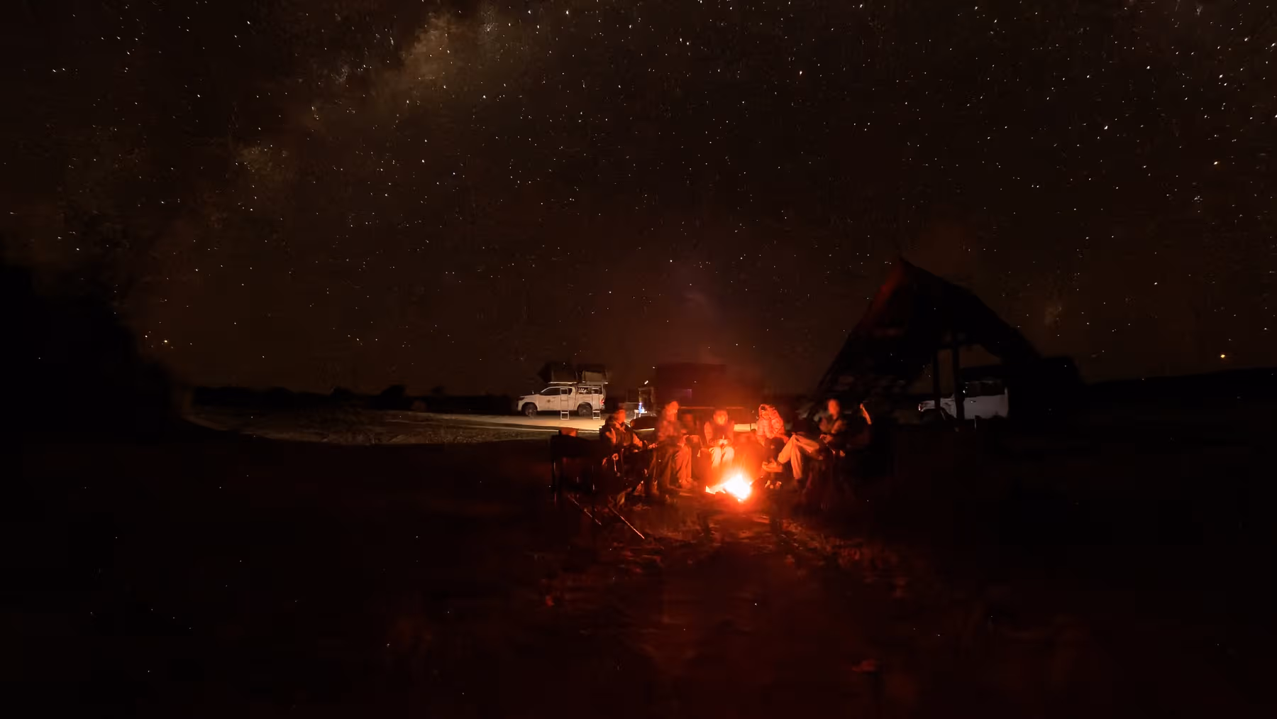 Mensen rond een kampvuur onder een heldere sterrenhemel met voertuigen en een tent in de achtergrond.