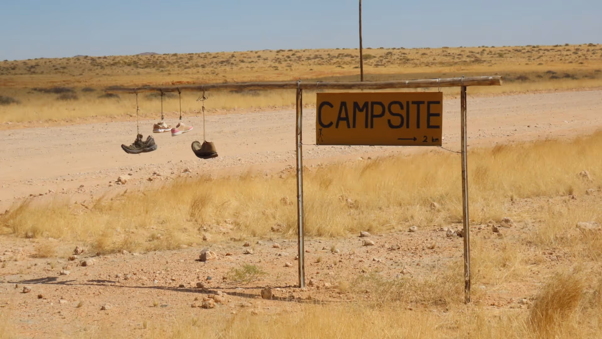 Bord langs een stoffige weg met tekst 'Campsite' en schoenen die aan een houten balk hangen in een droog graslandschap.
