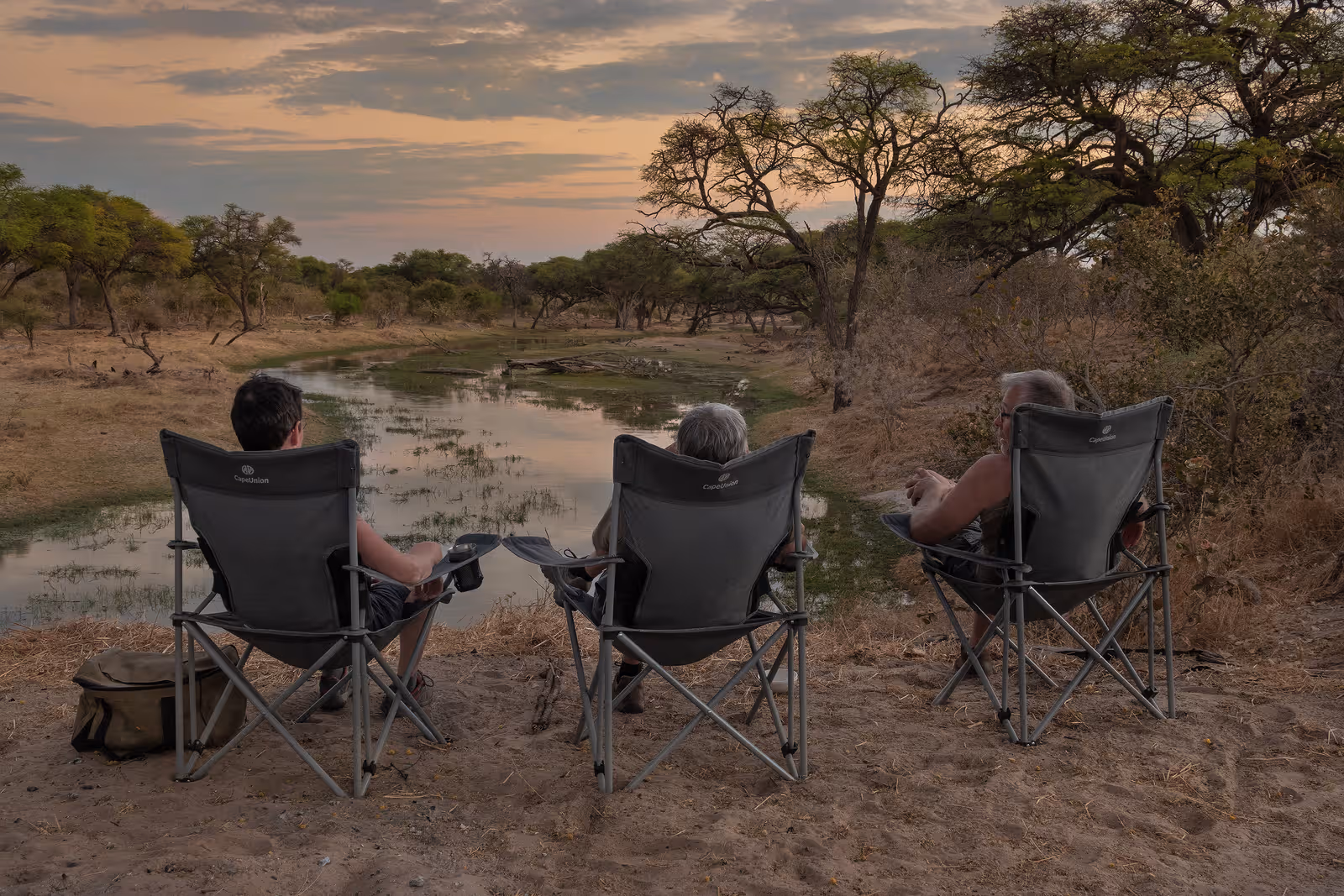 Drie personen zitten in campingstoelen aan een rivier tijdens de zonsondergang, omringd door droge bomen en struiken.