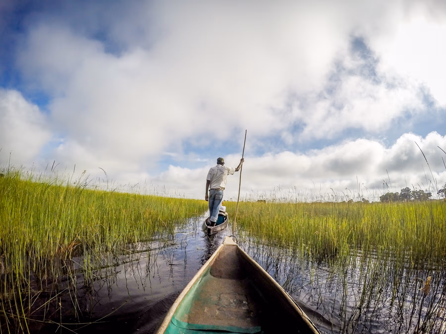Man met een stok staat in een smalle kano die door het water met groen gras aan weerszijden vaart onder een bewolkte blauwe lucht.
