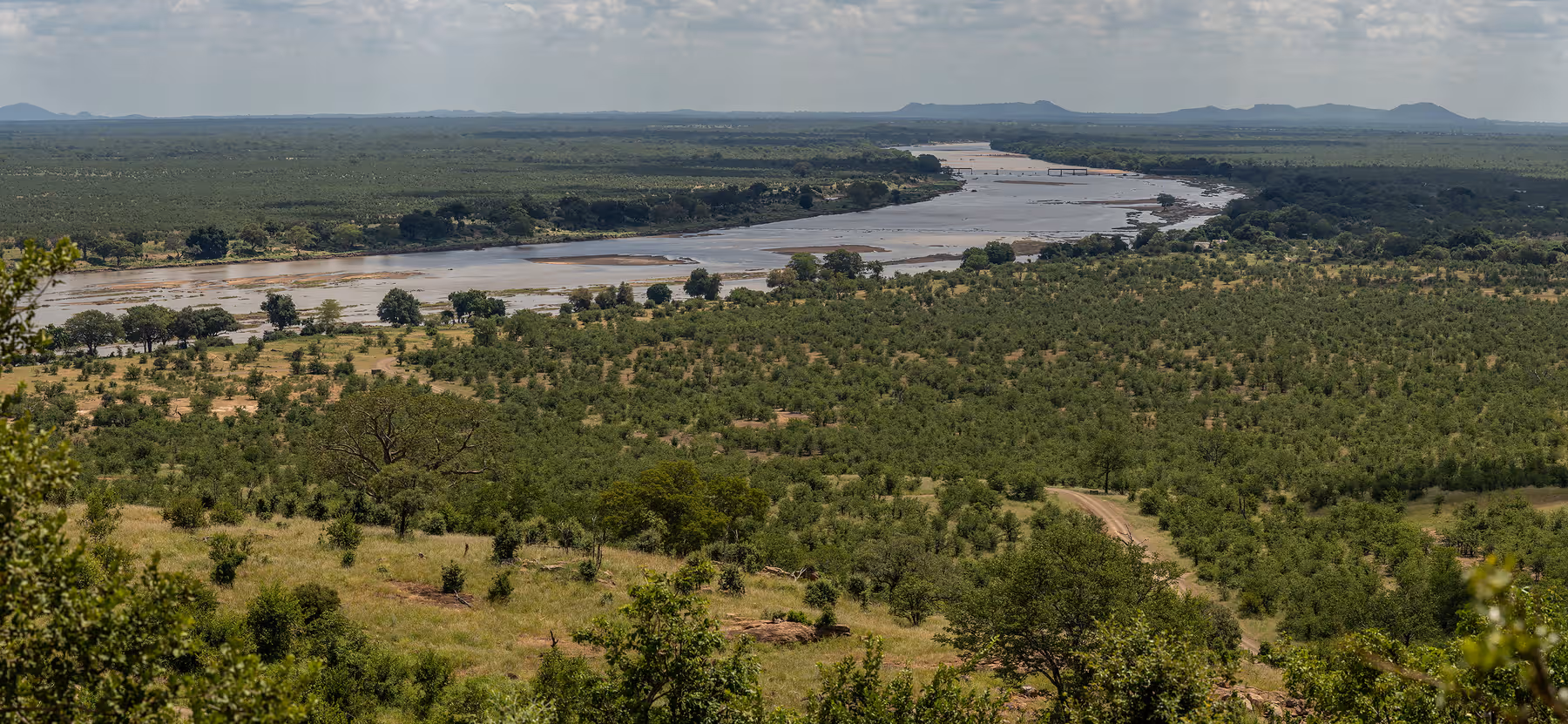 Uitzicht over een uitgestrekt landschap met dichte groene begroeiing, een brede rivier en bergen aan de horizon onder een bewolkte hemel.