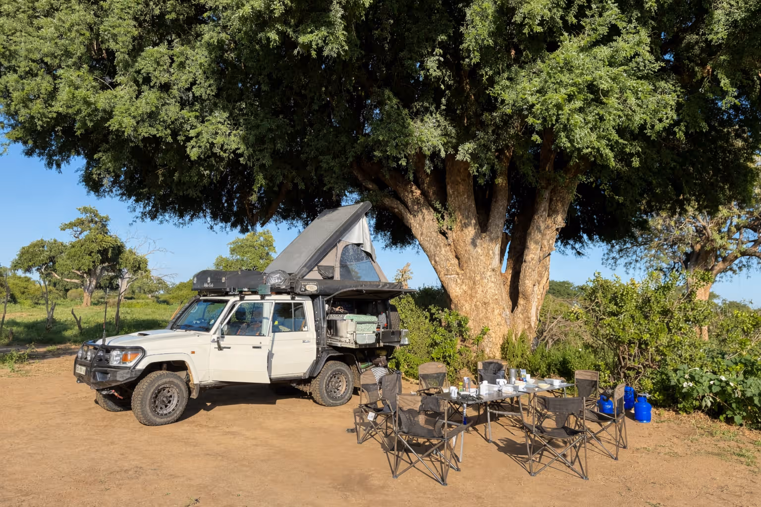 Een terreinwagen met een geopend daktentje geparkeerd onder een grote boom naast een campingtafel met zes vouwstoelen en kampeeruitrusting.