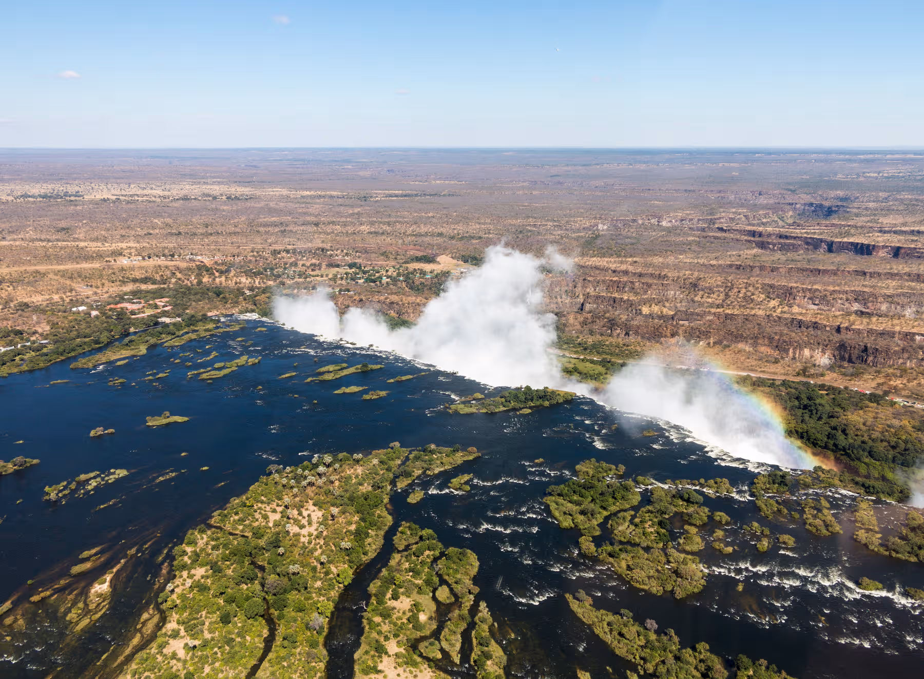 Luchtfoto van de Victoriawatervallen met een regenboog in de mist en de omliggende rivier en rotsachtige landschappen.