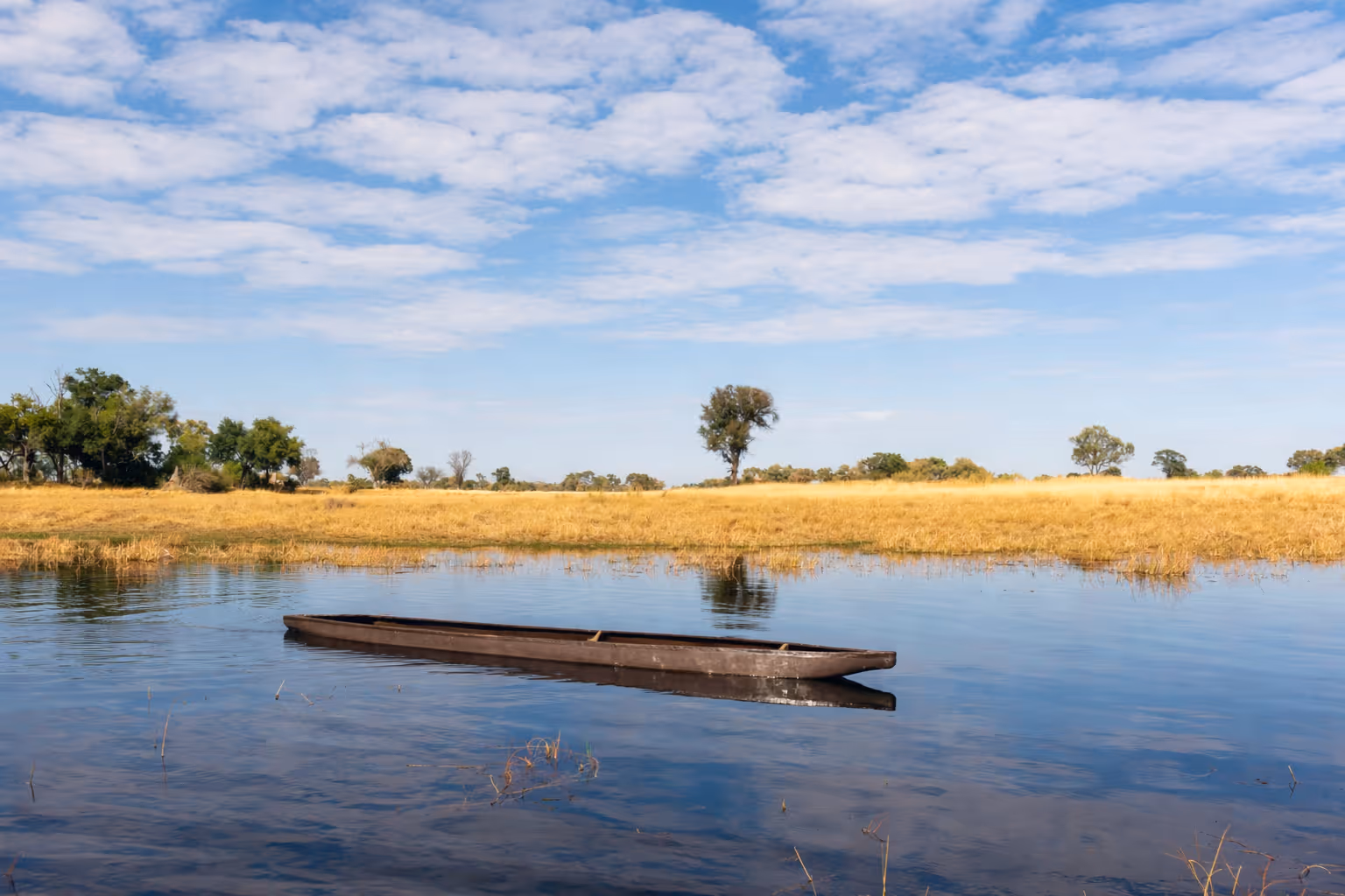 Mokoro drijvend op kalm water bij droog grasland met verspreide bomen onder een deels bewolkte hemel.