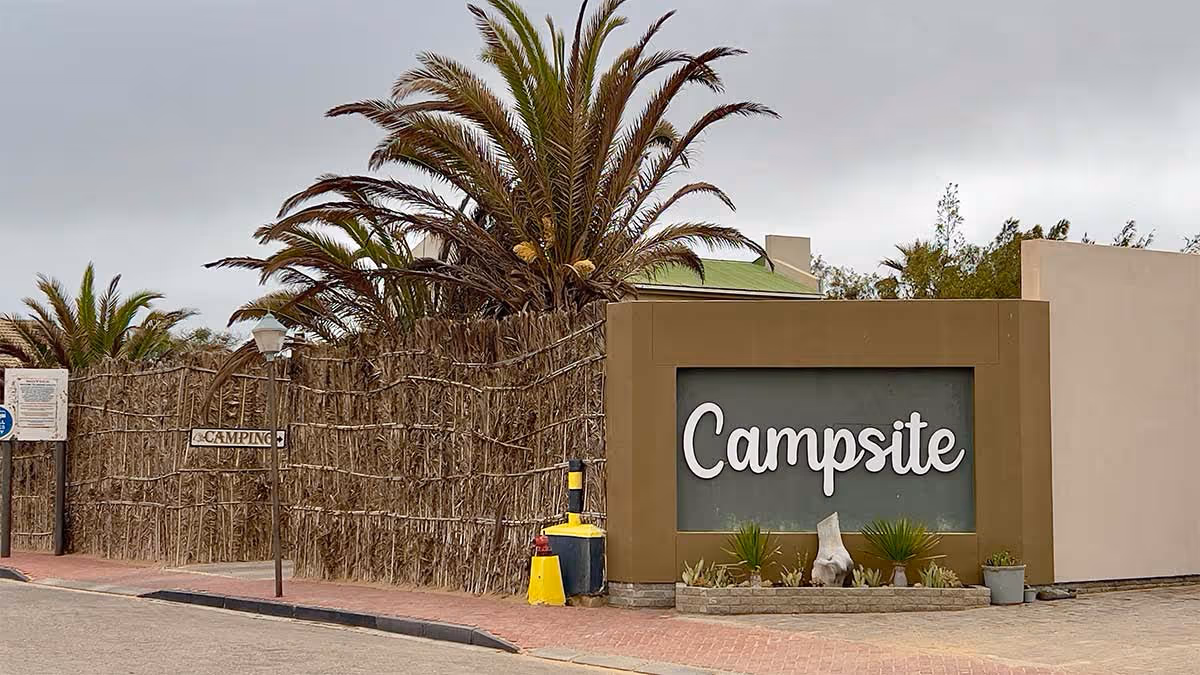 Ingang van een camping omgeven door palmbomen en een natuurlijke heg met een groot bord met 'Campsite'.