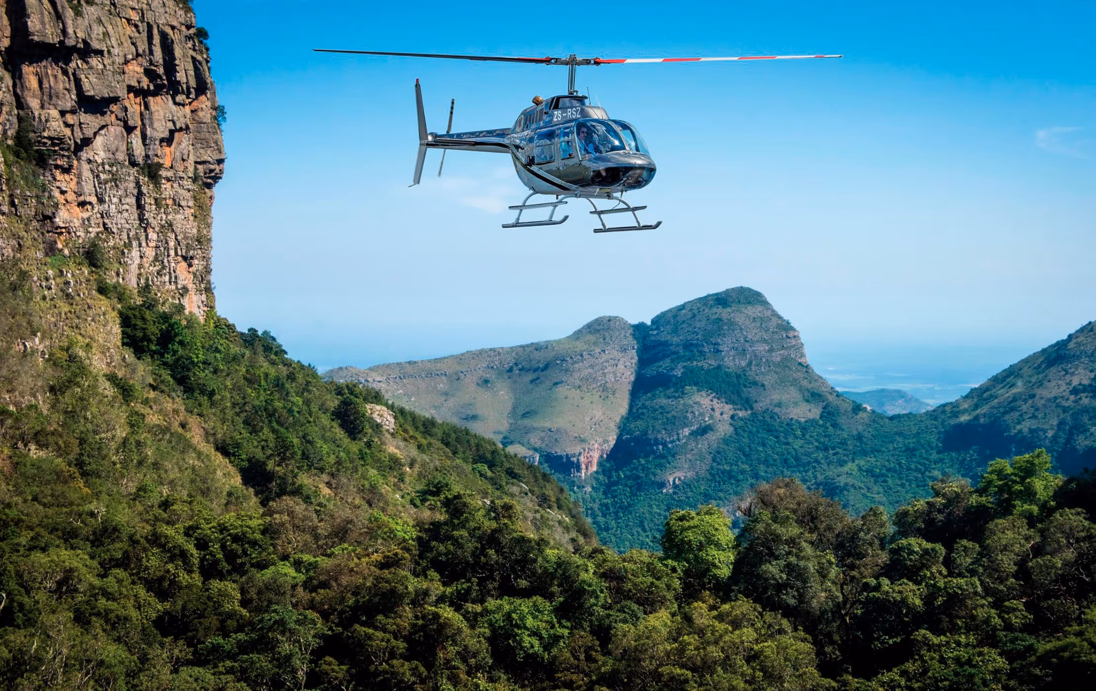 Helikopter vliegt boven een groen bebost bergachtig landschap onder een blauwe hemel.
