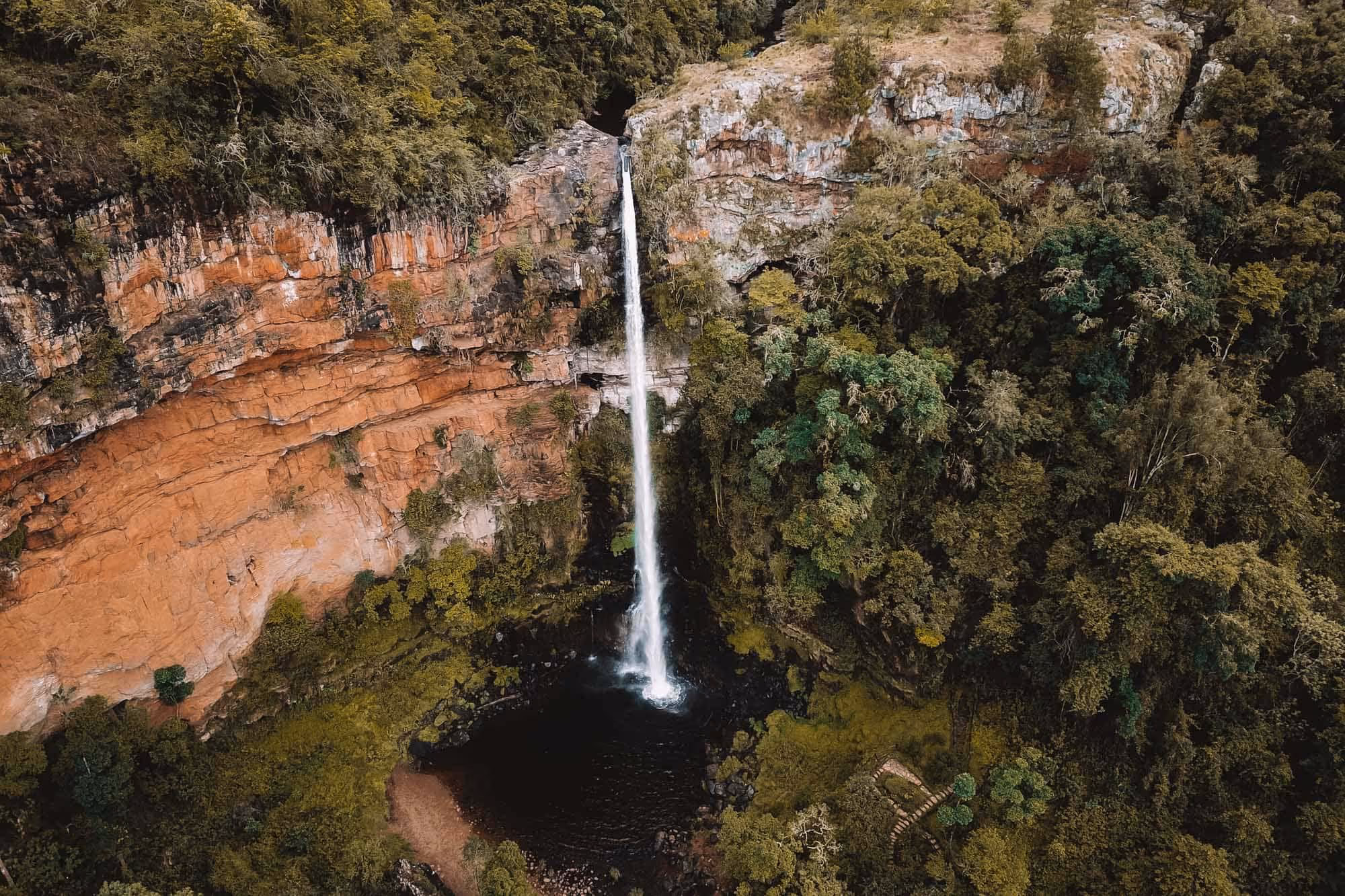 Luchtfoto van een smalle waterval die van een roodachtige rotswand naar een donker waterbekken in een dicht bebost gebied stroomt.