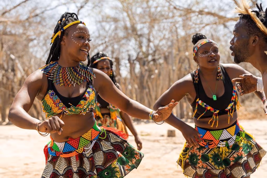 Mensen in kleurrijke traditionele Afrikaanse kleding dansen samen buiten in een droge, bosrijke omgeving.