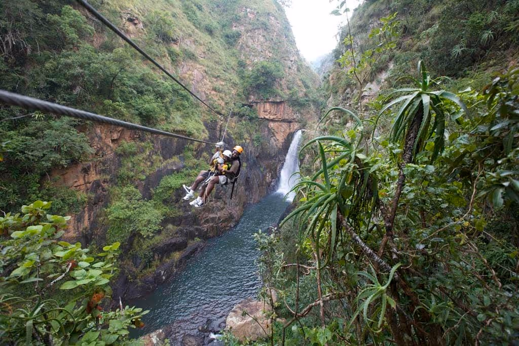Twee mensen aan een zipline boven een rivier met een waterval en groene rotsachtige heuvels.