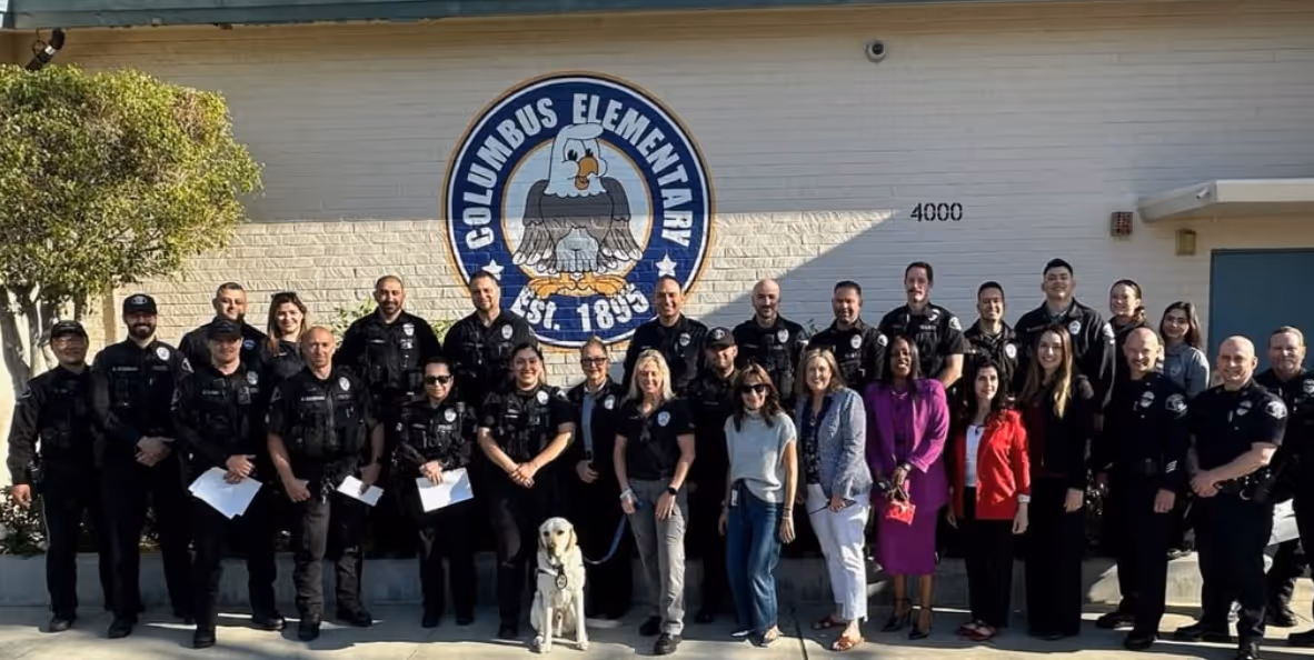 Group photo of police officers, staff, and a police dog standing in front of a Columbus Elementary School wall with an eagle logo.